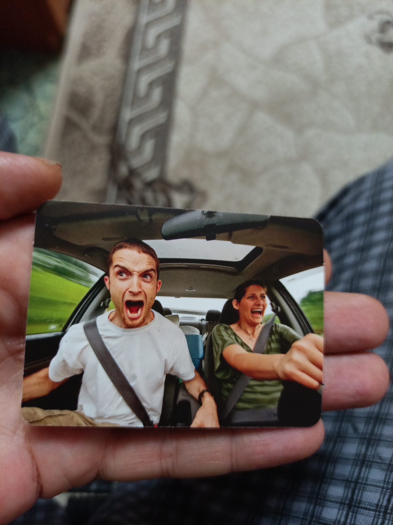 A small, rectangular photograph is held by a pair of hands in the foreground. The photograph depicts two individuals inside a car, both with mouths open and appearing to be screaming or laughing with extreme excitement or fear. The driver, a woman wearing a green striped shirt, has her hands on the steering wheel and is looking forward. The passenger, a man in a white t-shirt, is looking towards the left side of the frame. Both have their seatbelts on. The background of the photograph shows a blurred green landscape, suggesting motion. The photograph is slightly creased and the background behind the hands is out of focus, showing fabric textures. There is no discernible information within the image that places it in the city of Nyagan, Russia.