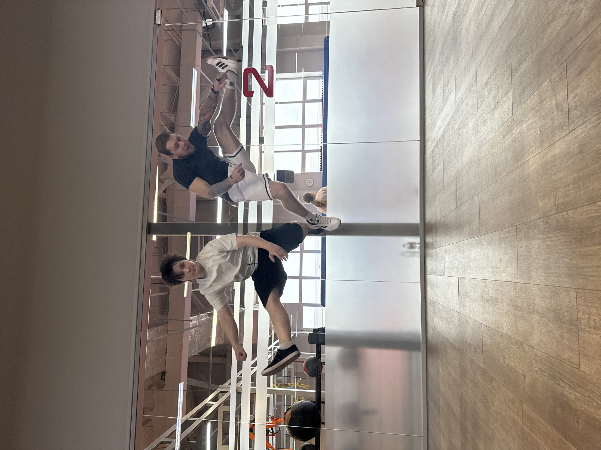 The image, if rotated 90 degrees counter-clockwise, depicts an indoor sports or fitness facility. A large, clear glass partition separates the viewer's space from the activity area.
Two adult males are visible through and reflected in the glass. One man, on the left side of the frame, is performing an inverted pose, possibly a handstand or a related acrobatic move, with his feet elevated. He wears a dark t-shirt, white athletic shorts with dark stripes, and white sneakers with black stripes. He has visible tattoos on his arms. The second man, positioned to the right, is in a dynamic, leaning posture, wearing a light-colored t-shirt, black athletic shorts, and black sneakers.
Behind the men, various gym equipment such as medicine balls and metal racks are partially visible through the glass. The floor on the photographer's side is a light-colored wooden floor with distinct planks.
The men appear to be engaged in physical training, possibly practicing acrobatics, calisthenics, or dynamic exercises. Their poses suggest intentional and controlled movements.
A large, stylized red letter "N" is prominently displayed on or behind the glass partition, appearing reversed due to reflection. The lighting is bright and even, characteristic of an indoor facility. The scene is located in Chaikovsky, Russia.