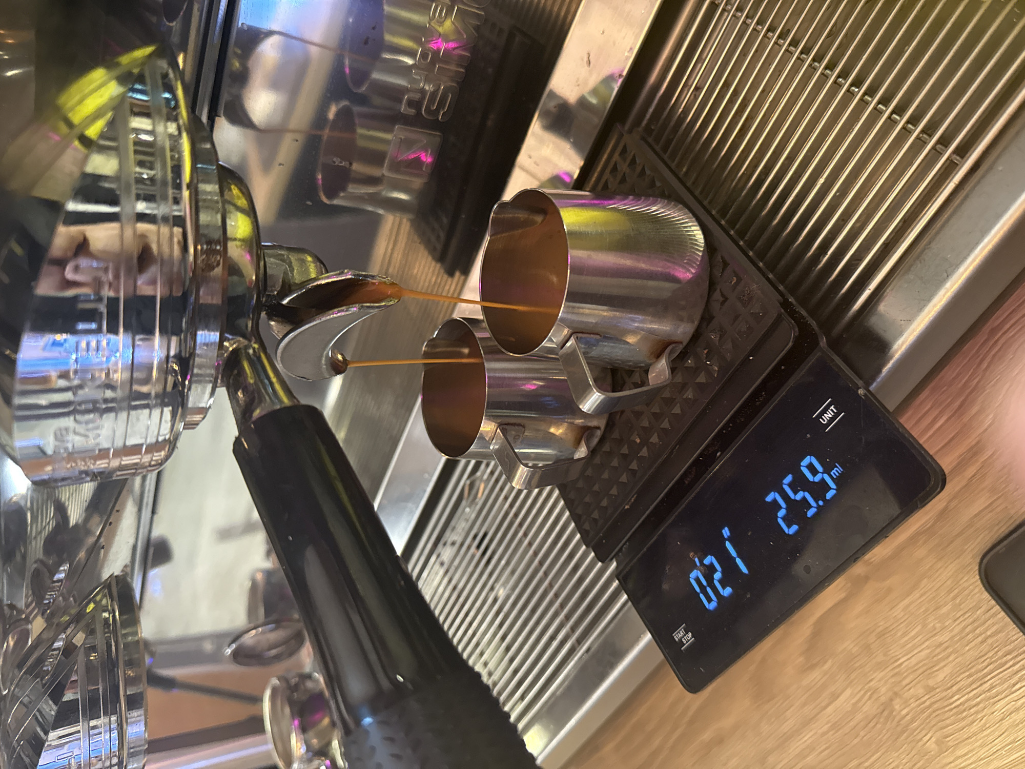 The image shows an espresso machine extracting espresso into two small metal cups. The cups are sitting on a digital scale that reads "25.9 ml". The espresso machine is stainless steel and reflects various colors in its surface. The background is a mix of other parts of the machine and a light-colored wooden surface. The overall setting appears to be indoors, likely a kitchen or cafe. There is no information in the image to suggest a location in Moscow, Russia.