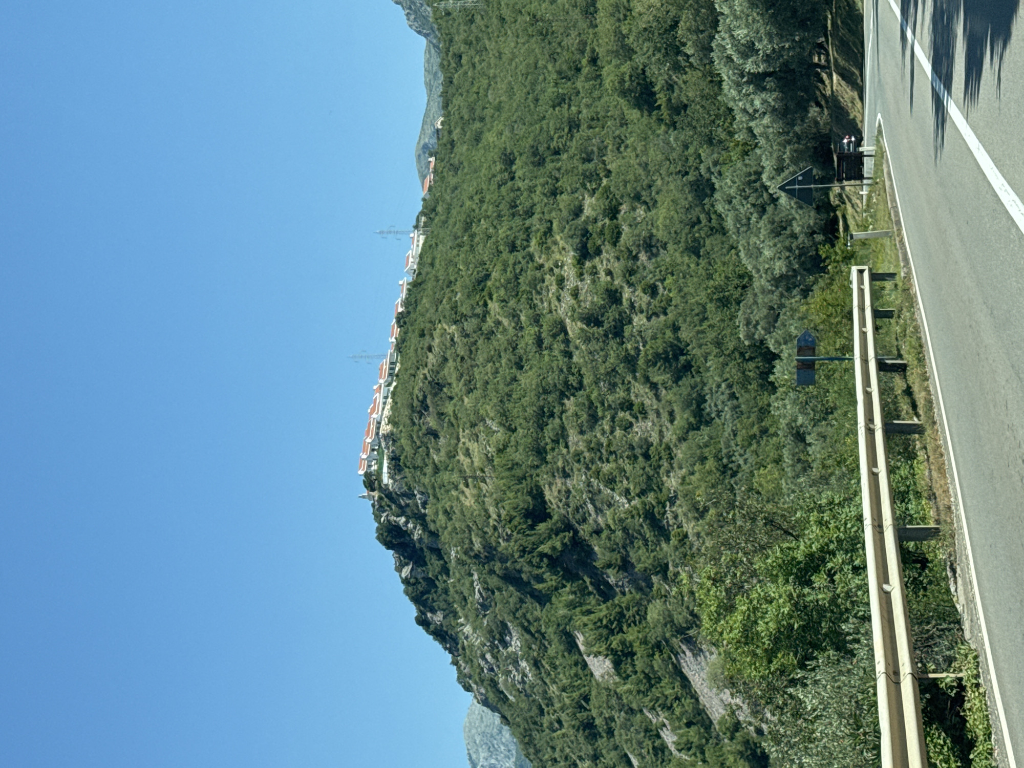 The image depicts a daytime scene in Blizikuće, Montenegro, showcasing a mountainous landscape with a road in the foreground. The upper third of the image is dominated by a clear, bright blue sky. A verdant, tree-covered mountain slope occupies the mid-ground and background, exhibiting rocky outcrops, particularly on the left side. Perched along the ridge of this mountain is a settlement composed of multiple white-walled buildings with distinctive red-tiled roofs. A prominent white structure, resembling a church or bell tower, is visible at the far left end of this elevated settlement. Several metal lattice power line towers are interspersed among the buildings and along the ridge. In the foreground, a paved asphalt road curves towards the right, featuring a solid white line marking its right edge. A light-colored metal guardrail runs along the left side of the road. Behind the guardrail, dense green foliage and trees extend up the hillside. On the right side of the road, more trees and vegetation line the curve. A blue directional sign with a white arrow pointing right is partially visible on the right shoulder, alongside a triangular warning sign. A dark-colored vehicle is visible in the distance on the right side of the road, partially obscured by the curve and foliage. No individuals are explicitly visible in the scene.