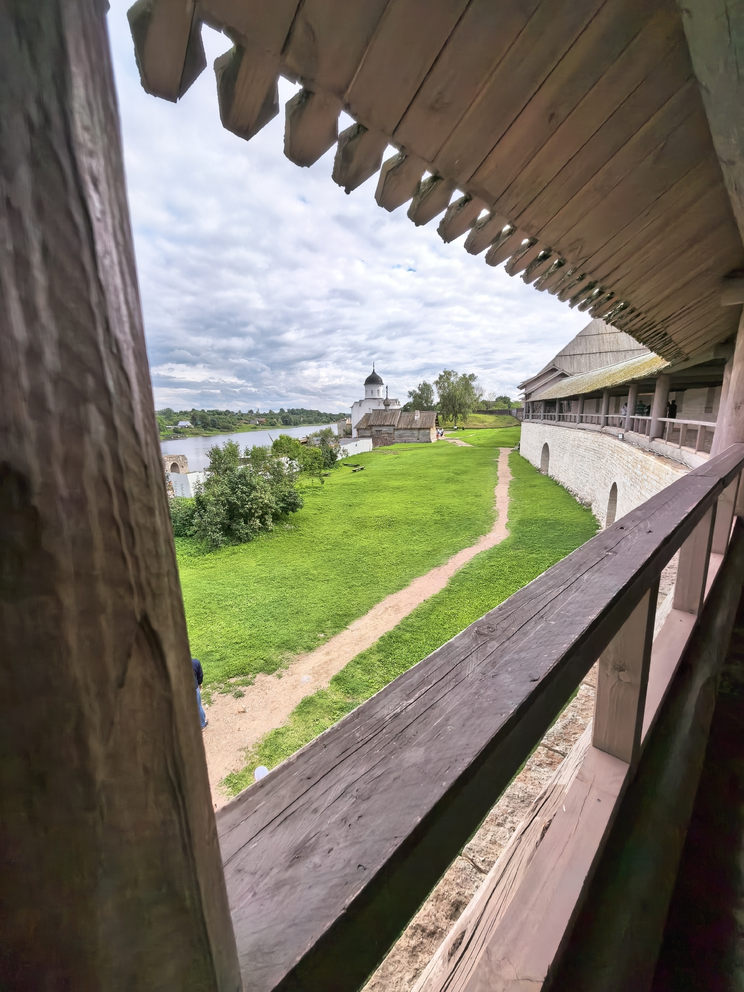 The image depicts an exterior view of the Staraya Ladoga fortress in Staraya Ladoga, Russia, looking out from a wooden structure. In the foreground, weathered wooden beams and railings form a framing element, suggesting a walkway or balcony. Beyond this structure, a large expanse of lush green grass stretches out, with a dirt path winding through it.
Dominating the middle ground is a light-colored stone wall, characteristic of fortress construction, with arched openings. Adjacent to this wall is a wooden building with a covered walkway supported by posts. Further in the distance, a white church with a dark dome and a smaller, dark-domed building are visible, along with some trees and greenery. A body of water, likely the Volkhov River, is present to the left, with a boat or barge on it. The sky is overcast with scattered clouds.
A single person, dressed in dark clothing and blue jeans, is partially visible on the grass path, walking away from the viewer. Another person, also dressed in dark clothing, stands on the covered walkway of the wooden building in the middle ground. The overall scene conveys a sense of historical architecture and a serene, somewhat overcast day.