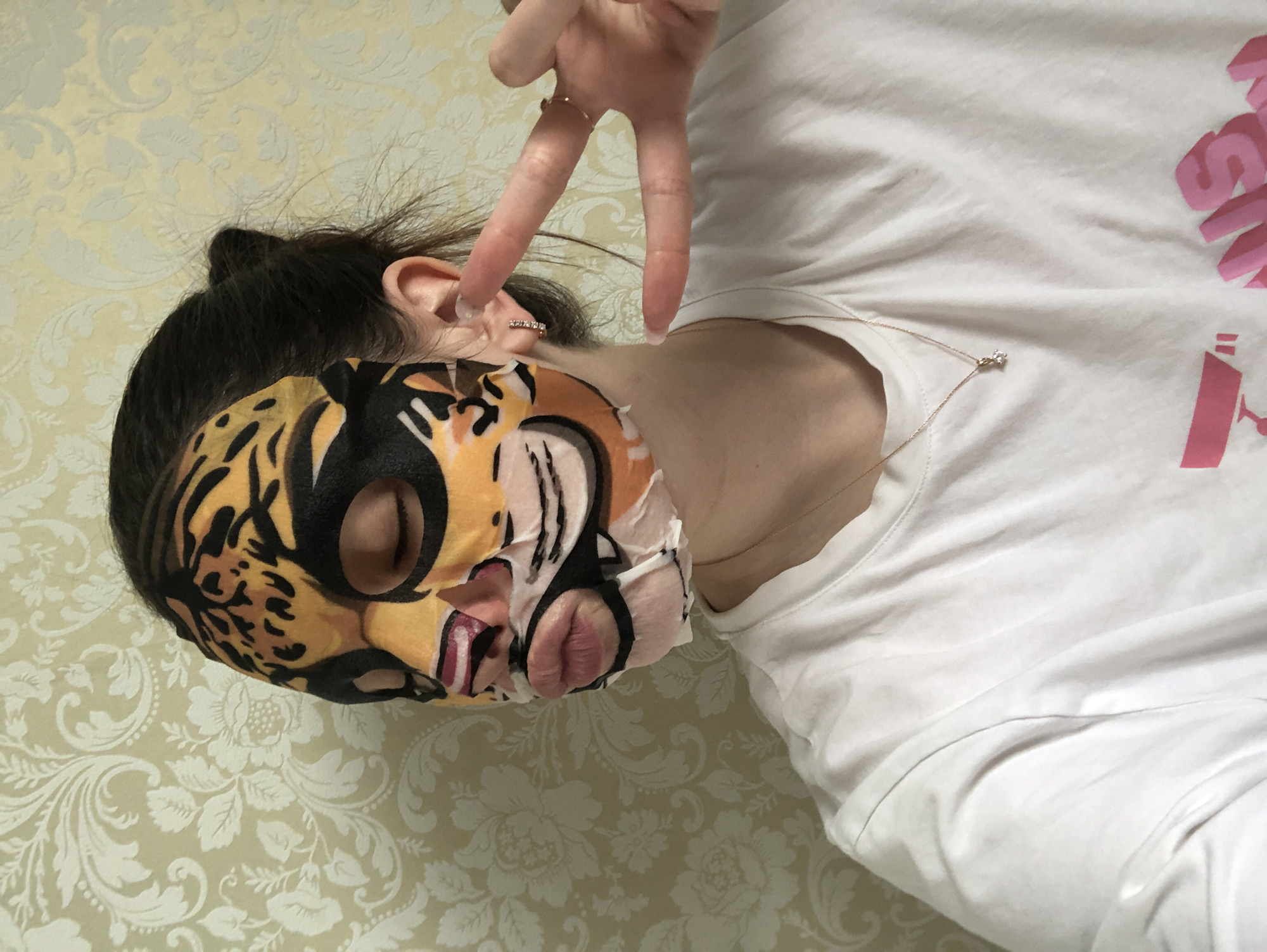 An indoor, vertically oriented close-up shot features a person wearing a playful tiger-faced sheet mask, presumably in Noviy, Russia. The individual is positioned against a light beige wallpaper adorned with a subtle, repeating white floral and damask pattern. The person's dark hair is pulled back, revealing an ear with a small, jeweled hoop earring. They are wearing a simple white short-sleeved t-shirt, on which pink graphic text, appearing to be Japanese Katakana, is visible on the right side of the chest. The visible characters spell out "メガドンキホーテ" which translates to "Mega Don Quijote." A thin, gold-colored necklace with a single, small, clear stone pendant rests on their collarbone. A hand, with nails painted with white tips, is raised to the upper right, forming a peace sign. A ring is visible on the index finger. The overall impression is casual and relaxed, with the sheet mask suggesting a skincare routine or a moment of self-care.