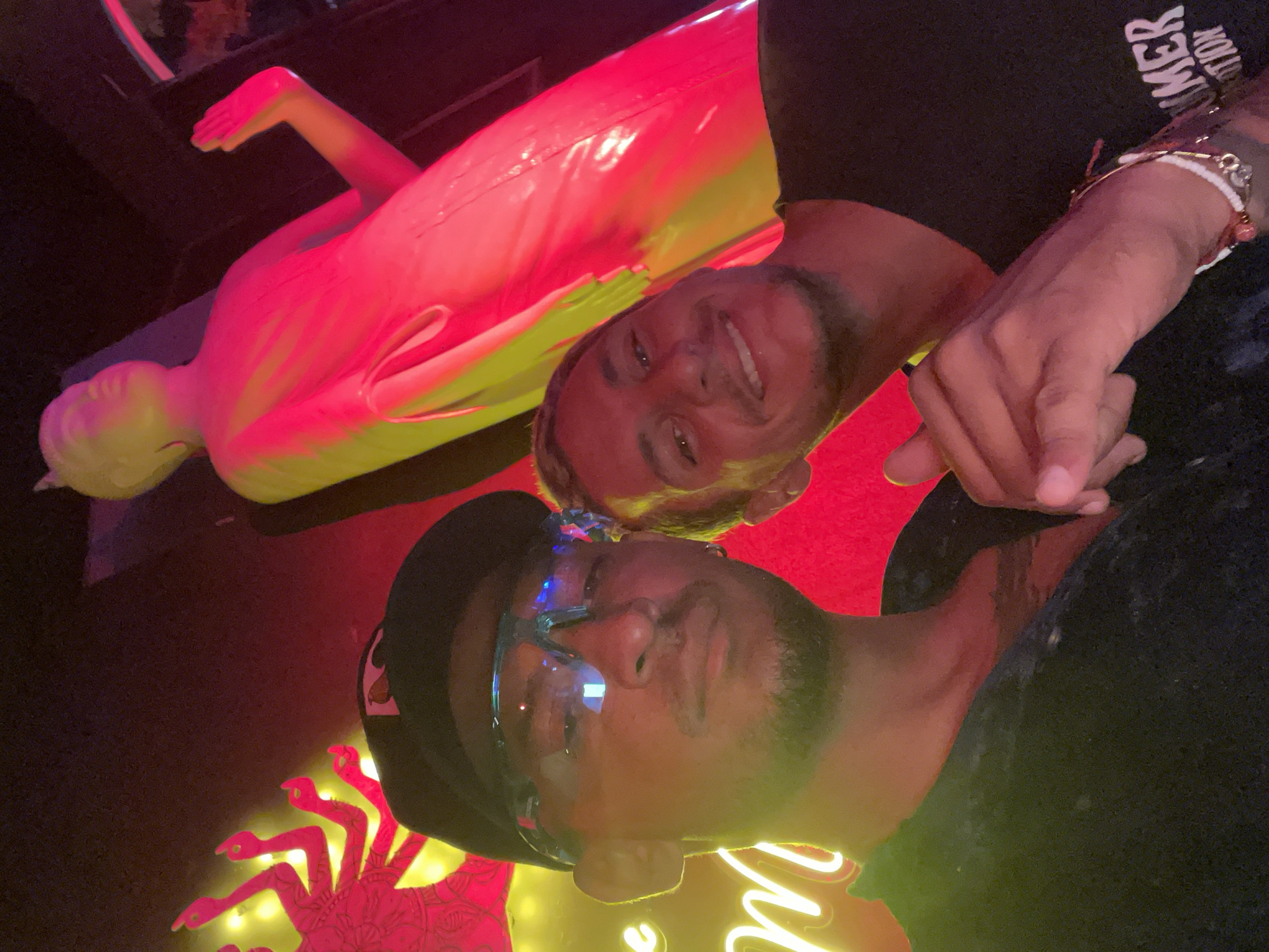 The image shows two men, one wearing glasses and a hat, standing next to each other in what appears to be a bar or club in Playa del Carmen, Mexico. The lighting is colorful, with red and yellow neon lights illuminating the scene. In the background, there is a mannequin lying down, as well as a neon crab decoration. The man on the left is wearing glasses and a hat, while the man on the right is smiling. The man on the right is wearing a black shirt and bracelets.