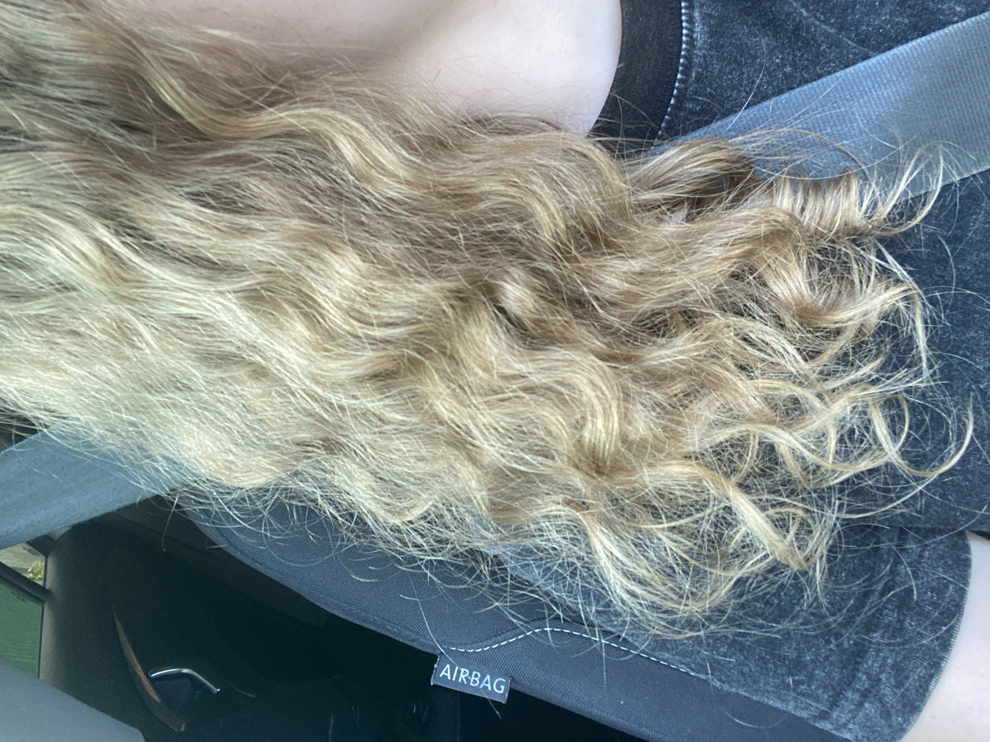 A close-up image shows a person's blonde, wavy hair in the foreground, with a portion of a dark gray car seat visible to the left. The car seat has a white stitched seam and a black label with white text that reads "AIRBAG". The background is blurred but appears to be the interior of a car with a green element visible through a window. The person is wearing a dark gray t-shirt. The scene is inside a car.