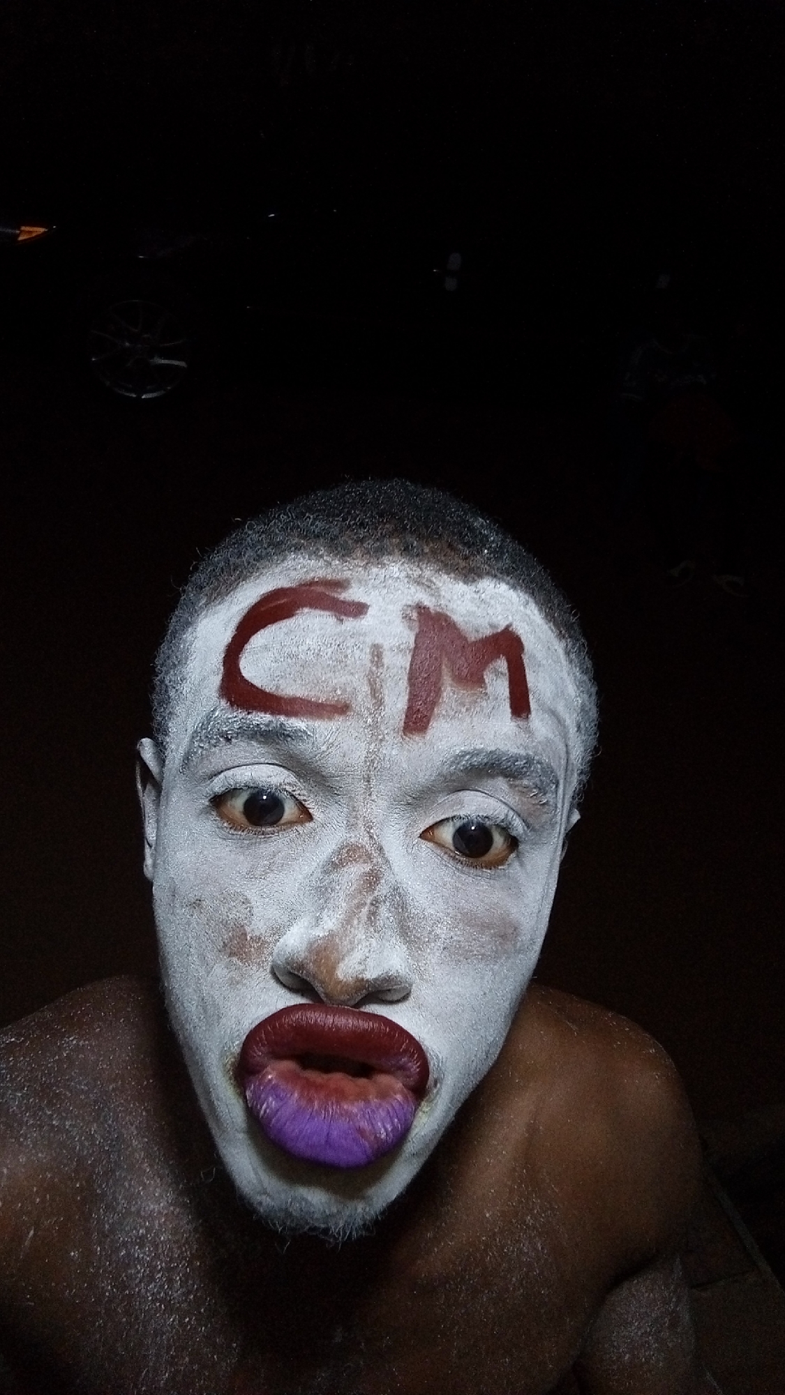 A close-up photograph shows a dark-skinned individual with white face paint. Red lettering "CM" is painted on the forehead. The lips are painted with a color gradient, transitioning from red to purple. The photo appears to be taken at night or in a dark environment, with a vehicle dimly visible in the upper part of the image. It is not possible to determine the location of the image as Onitsha, Nigeria without additional information.