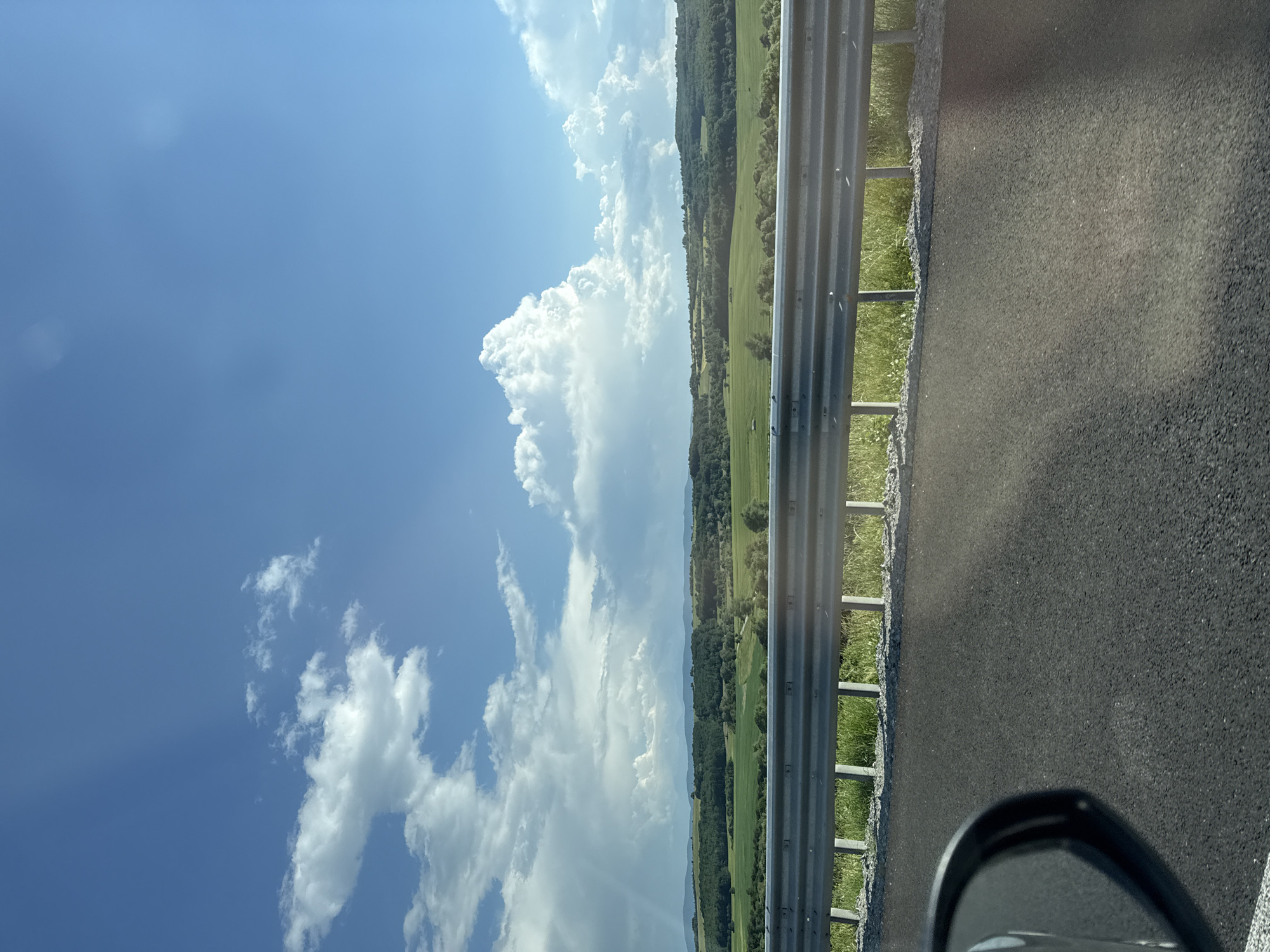 A scenic view of rolling green hills and a blue sky with scattered white clouds is captured from a roadside perspective. In the foreground, a dark asphalt road stretches across the lower portion of the image, with a portion of a car's side mirror visible on the left. A metal guardrail with vertical supports runs horizontally across the mid-ground, separating the road from the landscape beyond. The landscape consists of fields of varying shades of green, interspersed with patches of trees and denser forests on the distant hills. The sky is a vibrant blue, with dramatic white cumulus clouds, some of which appear to be building into storm clouds on the left side of the frame. The overall impression is of a rural or suburban environment on a clear, possibly humid day. The location is identified as Vakarel, Bulgaria.
