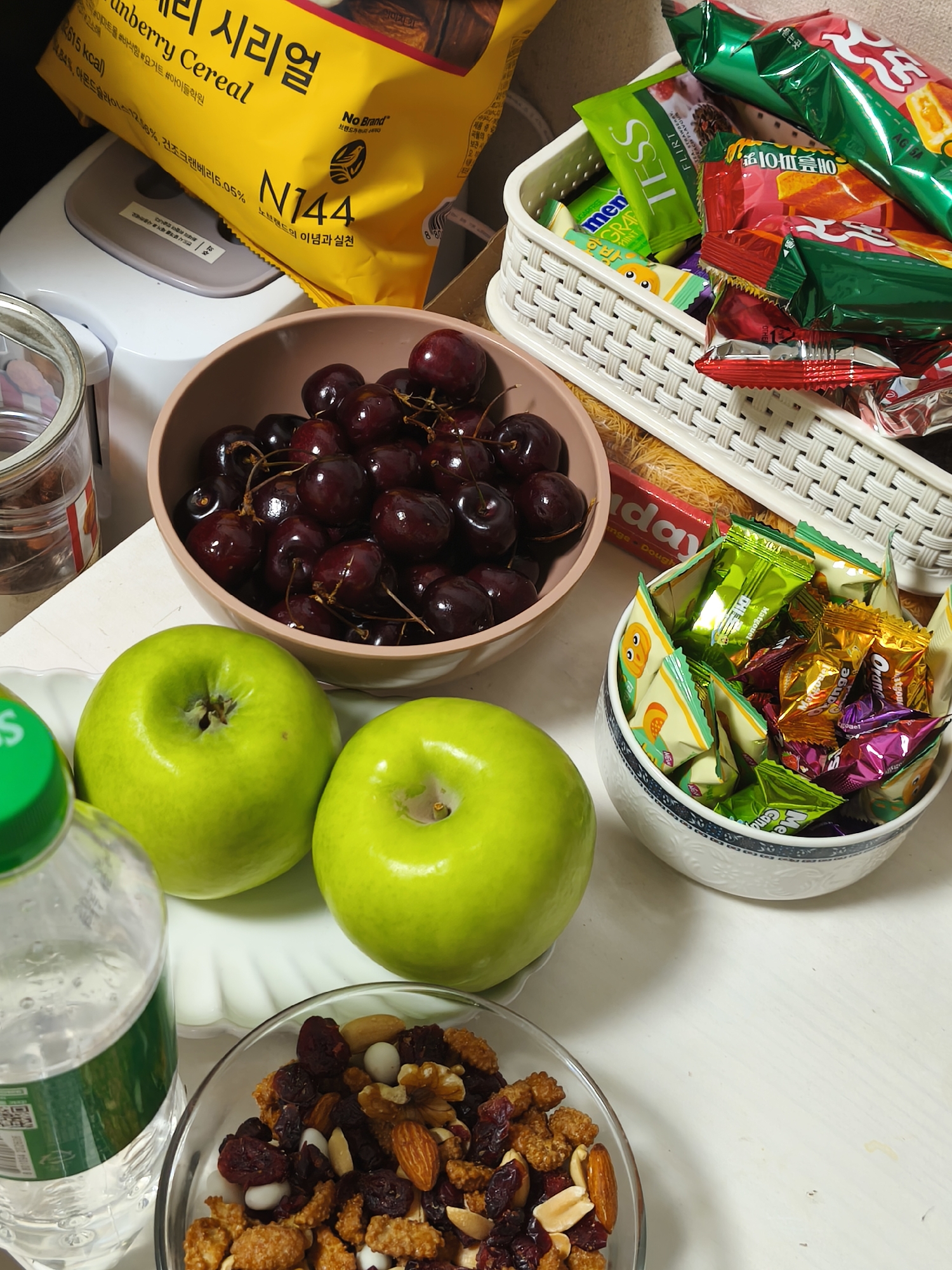 The image depicts a collection of snacks and fruits arranged on a white surface, likely a table. In the foreground, a clear glass bowl contains a mixture of nuts, dried cranberries, and what appear to be sweetened cereal clusters. To the left of the bowl, a partially visible plastic water bottle with a green label is present. Two bright green apples are positioned in the center-left of the frame, one slightly behind the other.
Behind the apples, a pink ceramic bowl is filled with dark red cherries. To the right of the pink bowl, a white woven basket holds an assortment of individually wrapped candies and snacks, many with colorful foil packaging. Some of these wrappers are green, red, and gold. A larger, red and white striped package of snacks is also partially visible in the basket.
In the upper left background, a portion of a yellow and black packaged cereal box with Korean text is visible, resting on top of a white appliance that resembles a bread maker. The overall environment suggests a casual setting, possibly a home or a snack station. The lighting is bright, indicating an indoor setting with artificial or natural light. No people are visible in the image. The scene is static and shows a collection of food items. The items are laid out on a white table. The image is taken from a slightly elevated perspective, looking down on the arrangement. The context of Asan-si, South Korea, is not directly inferable from the visual content of the image itself, as there are no identifying landmarks or cultural markers present.