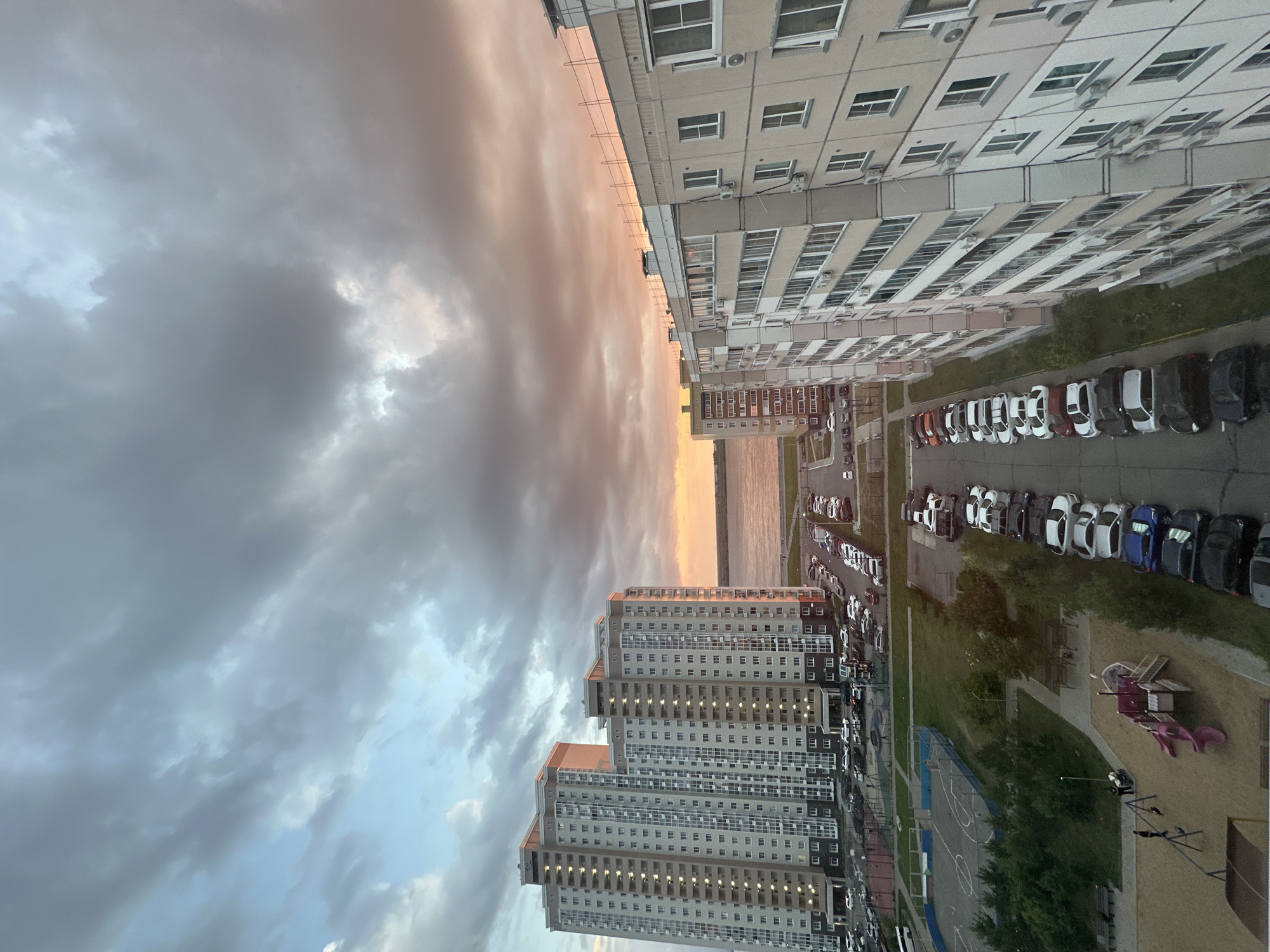 The image depicts a cityscape at sunset, likely in Khabarovsk, Russia, characterized by high-rise residential buildings and a body of water in the distance. The sky is filled with dramatic clouds, with a gradient of colors from light blue to pink and orange, indicating the sun is setting.
In the foreground and midground, several multi-story apartment buildings are visible. These buildings are modern in design, featuring numerous windows and balconies. They are constructed with light-colored facades, possibly concrete or stucco. The buildings are densely packed, with parking lots and green spaces interspersed between them.
A significant portion of the image is dedicated to a long, parallel row of parked cars along a road or parking lot. The vehicles are of various makes and models, predominantly passenger cars, and are parked in an organized manner. The cars are a mix of colors, including black, white, silver, and blue.
Further into the scene, there is a sports field, possibly a basketball or soccer court, with fencing around it. Adjacent to the sports field is a children's playground, featuring a slide and swings, with sand on the ground. Two adults are present near the playground, one pushing a child on a swing, and another adult standing with a stroller.
In the background, a wide river or bay is visible, with a distant shoreline. The overall impression is one of a residential urban environment during the evening hours, with the sunset creating a visually striking backdrop.