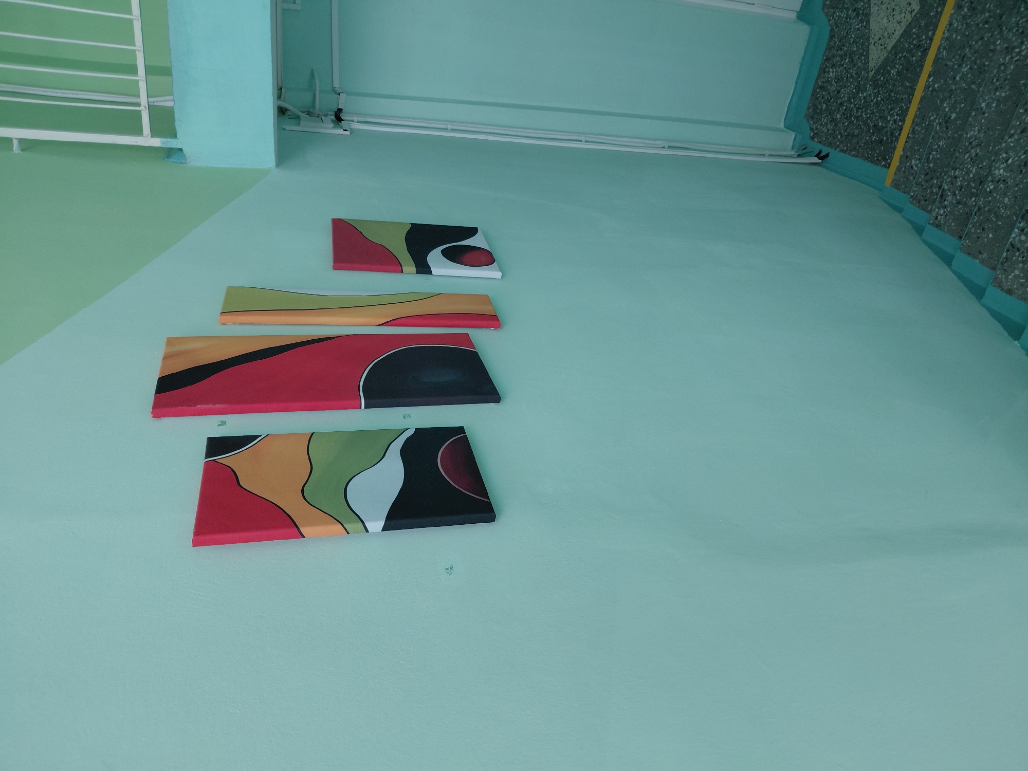 The image captures an indoor architectural scene, likely a hallway or stairwell in a public building in Nadym, Russia. The walls are painted in a dominant light teal or mint green, with a portion in the top left showing a lighter green shade that forms a sharp, geometric diagonal line where it meets the main teal color.
Four abstract, rectangular artworks are mounted on the teal wall, arranged vertically in a slightly staggered progression from top to bottom. Each painting features flowing, organic shapes filled with a vibrant color palette including red, black, yellow, green, orange, and white.
In the upper left, a white metal railing is visible, suggesting a balcony, upper landing, or staircase. Along the top of the wall, several white pipes or conduits run horizontally. The floor, visible in the upper and lower right sections, is made of a speckled, dark gray material, possibly terrazzo, with a prominent yellow stripe marking an edge, indicative of a stairwell.
The scene is devoid of people or any ongoing activity, presenting a static view of the building's interior. The lighting is even, suggesting artificial illumination or diffuse natural light. There is no discernible text in the image. The overall aesthetic is clean and functional, with the abstract paintings adding a splash of color and modern design.