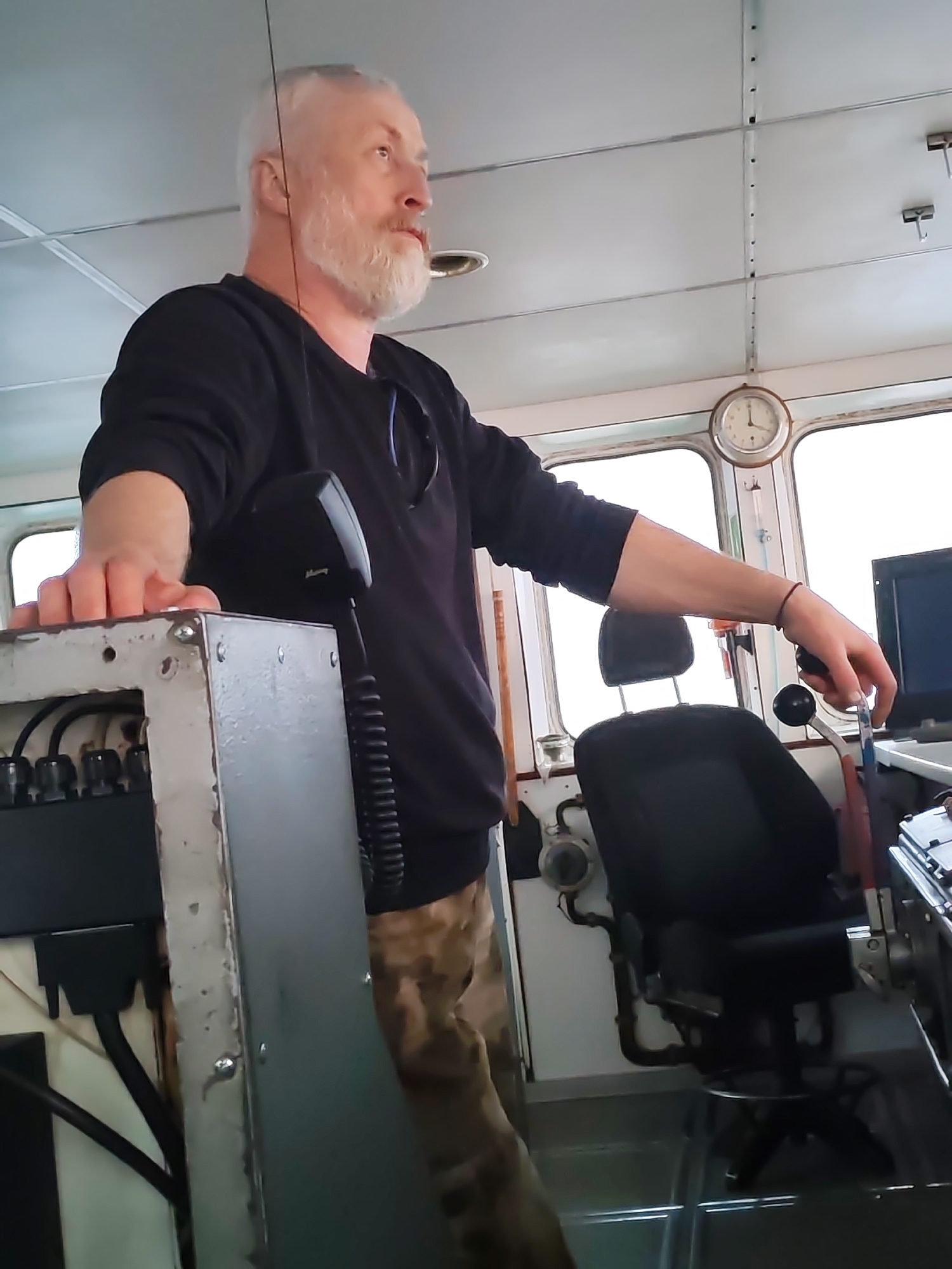 The image depicts a man with grey hair and a beard in what appears to be the bridge of a vessel. The man is wearing a long-sleeved black shirt and camouflage trousers. He is standing with his left hand resting on a grey control panel and his right hand operating a lever. In the background, there is a clock on the wall and a black chair. The surroundings suggest an enclosed space with windows. There is no explicit information about the location in the city of Kakkarovo, Russia.