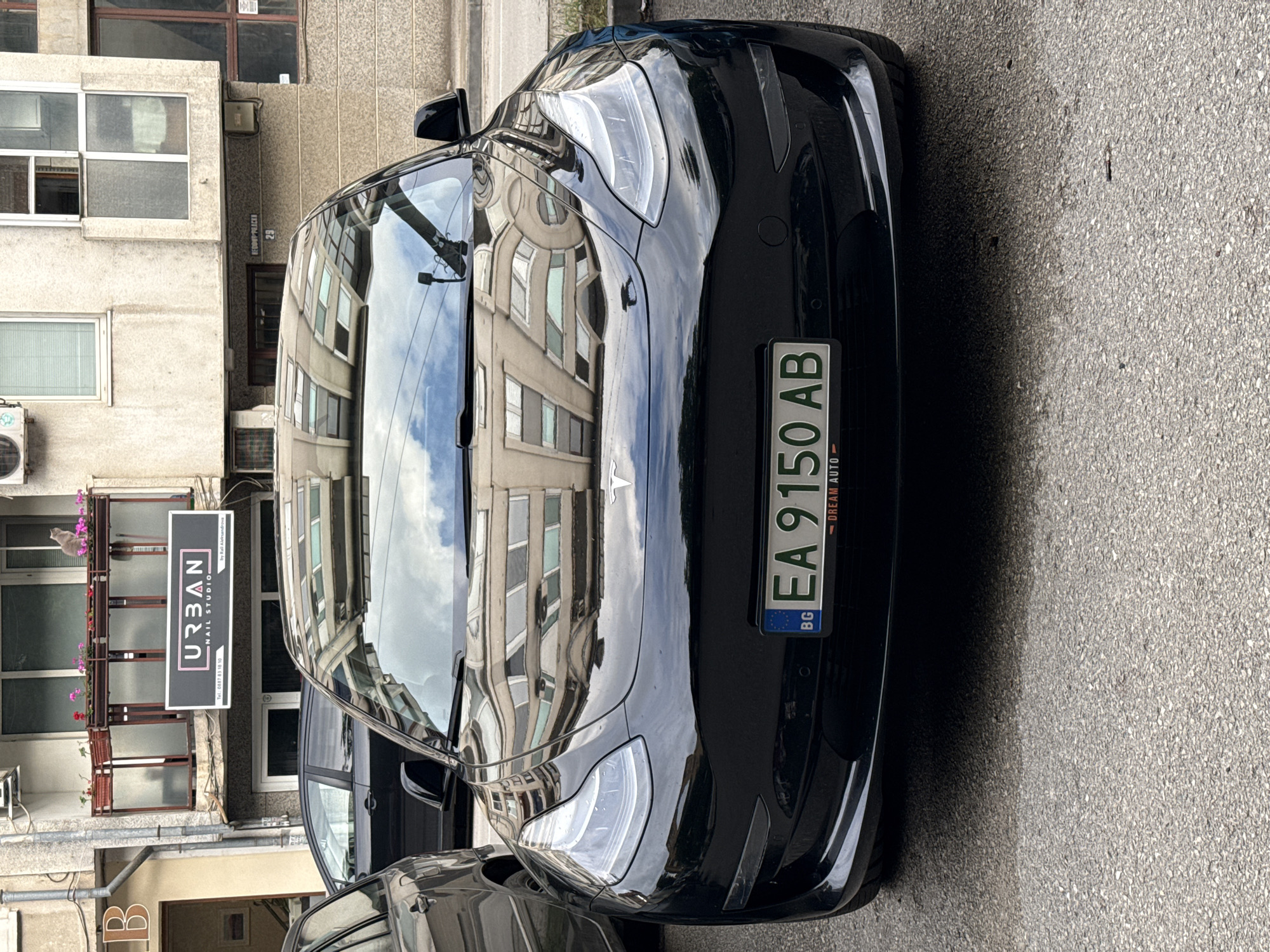 This image depicts a black Tesla Model 3 parked on an urban street in Pleven, Bulgaria, during what appears to be daytime with an overcast or partly cloudy sky. The car is positioned at an angle, showcasing its front and passenger side. The highly reflective surface of the car, especially the hood and windshield, mirrors the surrounding multi-story buildings and the sky above.
The vehicle displays a Bulgarian license plate with the registration "EA 9150 AB", accompanied by "BG" and "DREAM AUTO" on the plate frame. The street features asphalt pavement.
To the left of the car, several buildings line the street. One building prominently displays a sign for "URBAN NAIL STUDIO by Rali Aleksandrov," including the contact number "Tel: 0887 83 18 10." Further along, a street sign is visible with the text "REDOUT PRACKE 29", indicating a specific address. A small 'B' is also visible on a building near the bottom left. Air conditioning units are mounted on the exterior of some buildings, and a small cat can be faintly seen on a balcony above the nail studio sign. The scene is static, with no people visible, offering a snapshot of a typical street environment.