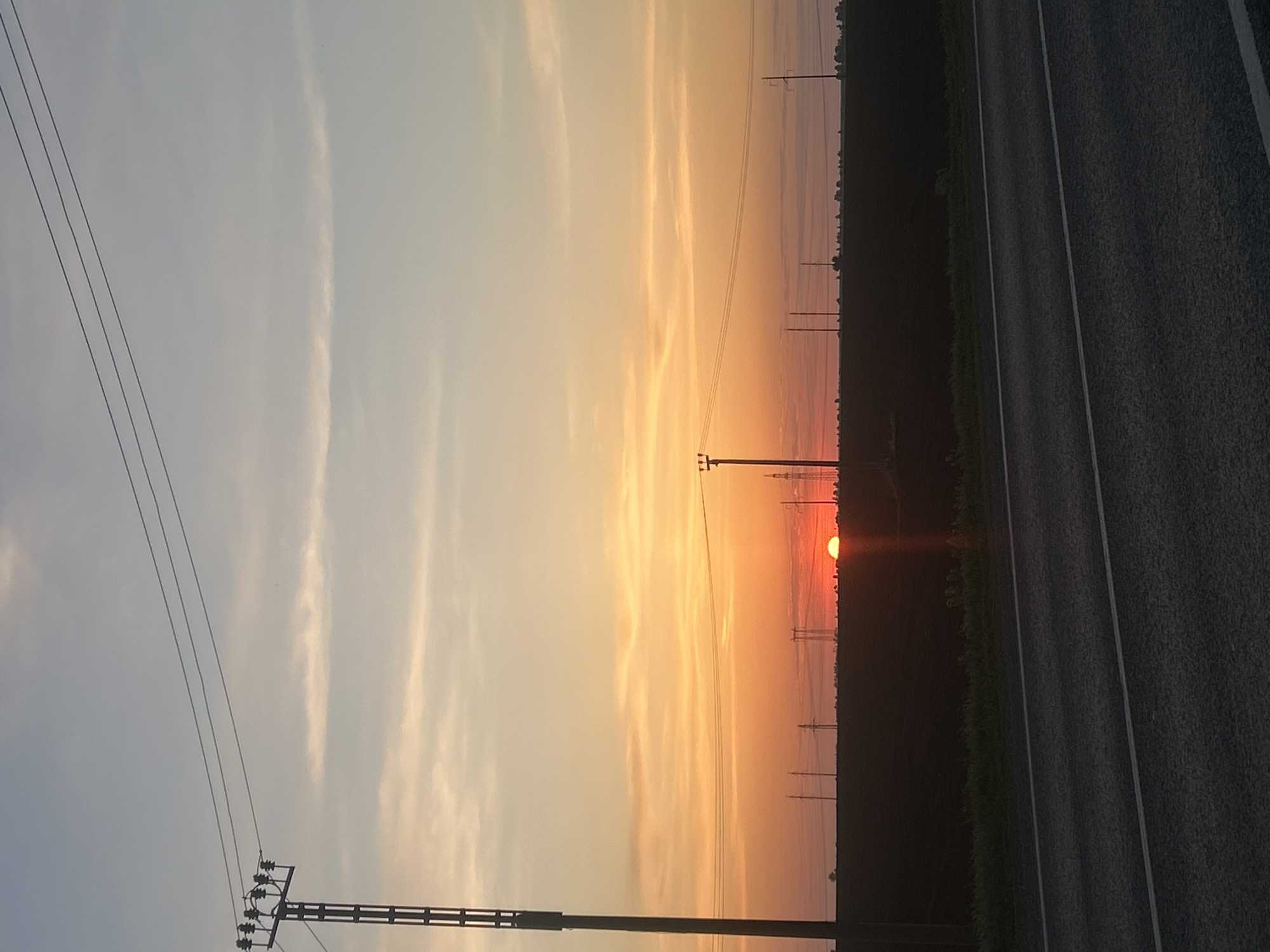 The image captures a sunset scene near a road in Uziukovo, Russia. The sky is a gradient of colors, ranging from pale blue to orange, with streaks of clouds. Power lines and utility poles extend across the frame. On the right side of the image, there is a dark, vegetated area adjacent to an asphalt road with painted lane markings. The sun is setting on the horizon, appearing as a reddish-orange orb.