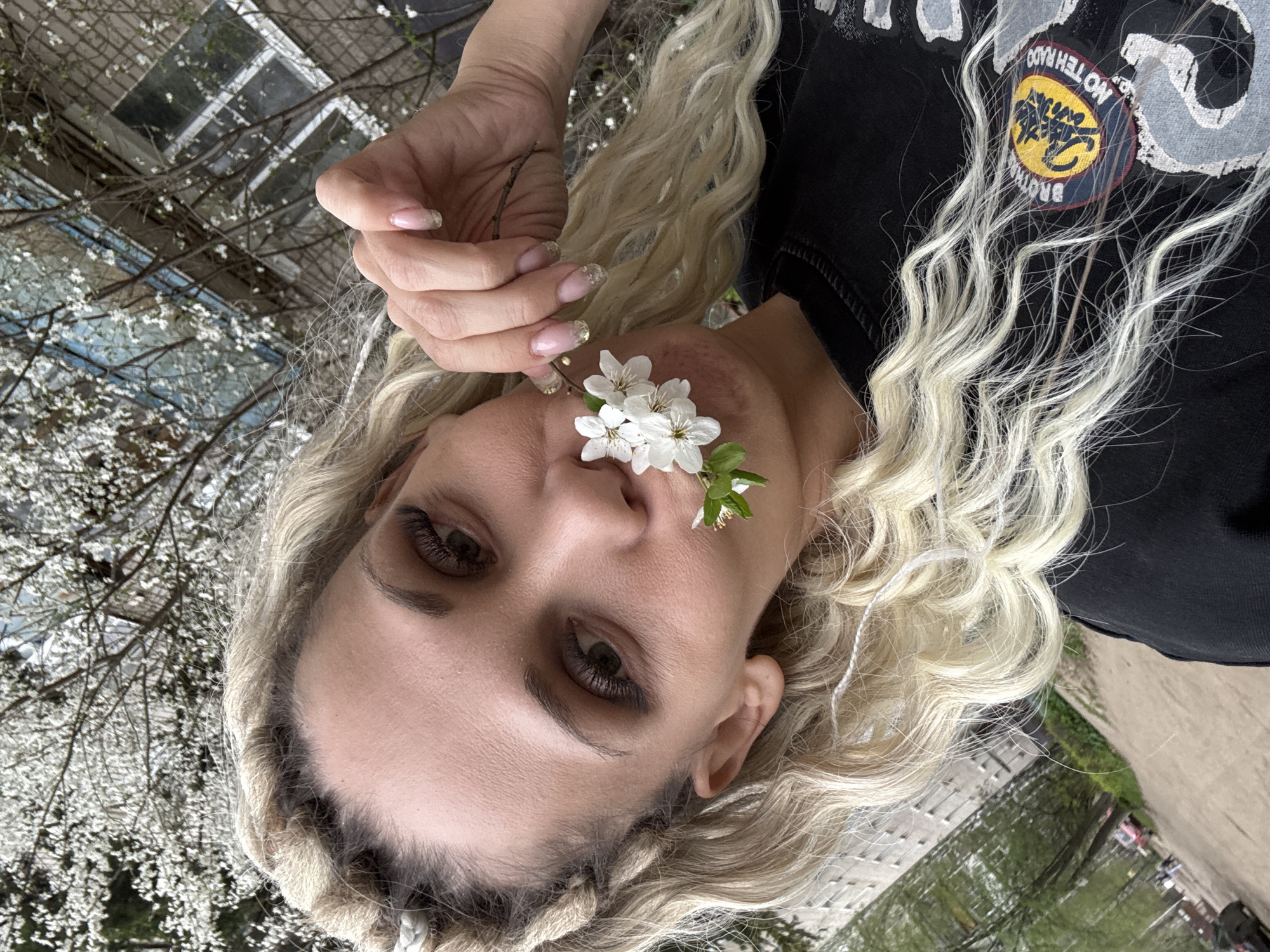 A woman with blonde braided hair and brown eyes is holding a small branch of white flowers to her nose. She is wearing a black shirt with a logo on it. The background includes a building and trees with white flowers. The location is Rusanovo, Russia.