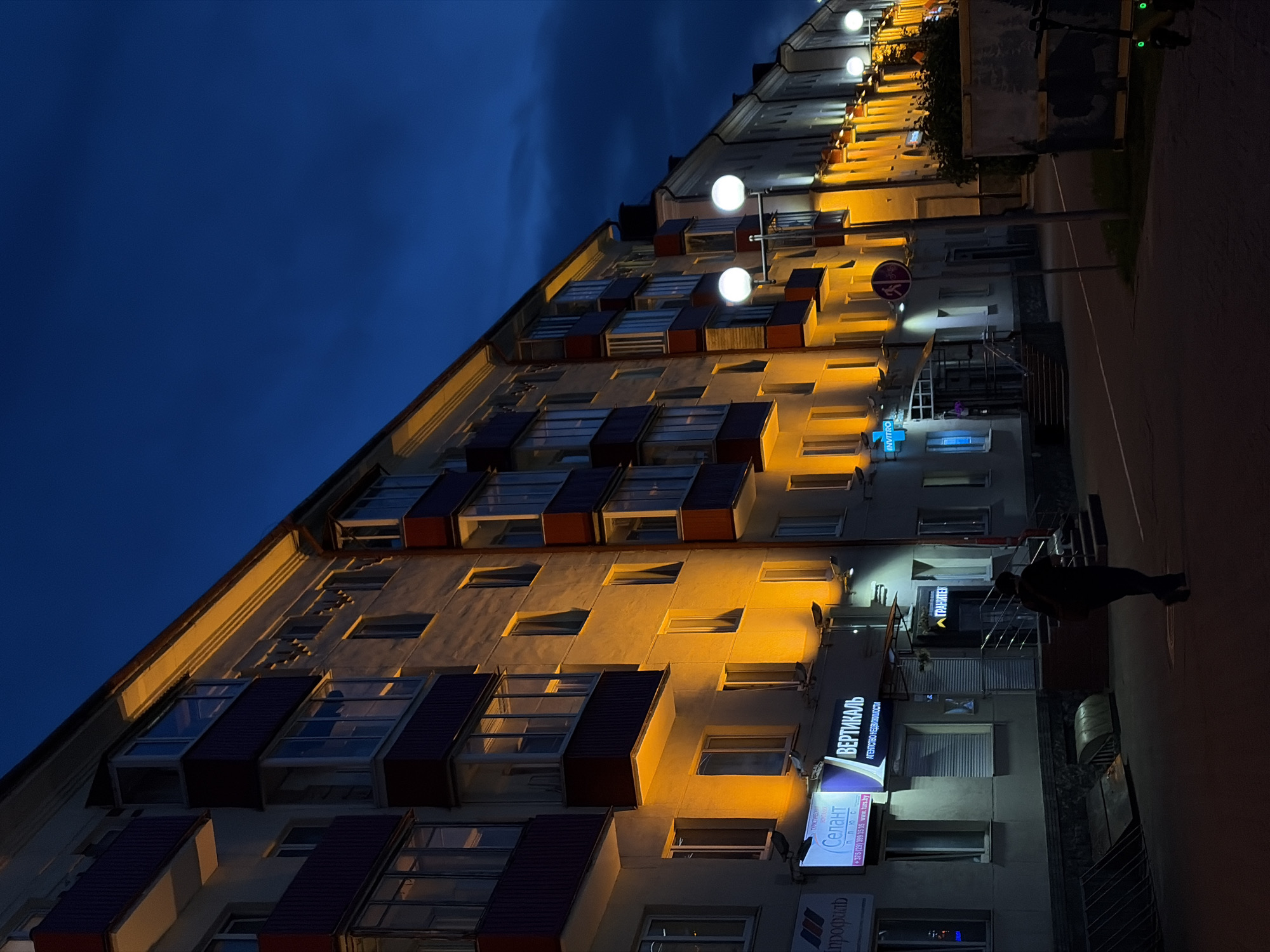 This night-time image captures a multi-story building along a street in Minsk, Belarus, under a dark blue sky. The building, viewed at an upward angle, dominates the left side of the frame, with its light-colored facade warmly illuminated by numerous yellow-orange streetlights mounted on its exterior. Many windows are visible, some with dark-colored awnings or window boxes.
On the right, a dark sidewalk or street runs alongside the building. A silhouetted individual is walking on this path in the lower right corner. Further up, partially visible bicycles or scooters are parked near a circular red-bordered traffic sign depicting a bicycle.
Several commercial signs are displayed on the lower levels of the building. These include a prominent blue sign for "INVITRO," a white sign for "ВЕРТИКАЛЬ АГЕНТСТВО НЕДВИЖИМОСТИ" (Vertikal Real Estate Agency), and another sign for "Селант плюс" (Selant Plus) with contact details: "+375 (29) 399 35 35 www.turs.by". Other visible texts include "ГРАНИТЕК" (Granitek) and a partial sign for "профиль" (profile). The scene depicts a quiet urban street at night.