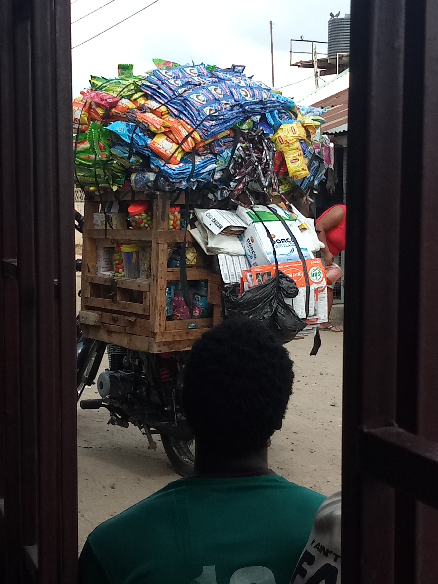 The media file shows a motorcycle heavily laden with goods, likely for transport or sale. The motorcycle is parked on a dirt road, and a person is standing in the foreground, looking away from the camera. The motorcycle's cargo consists of numerous packets of what appear to be snacks or detergents, stacked high and secured with ropes. A wooden crate is attached to the motorcycle, holding smaller items like candies and packaged goods.
In the background, a woman in a red top is visible, bending over some items. There are also buildings with corrugated roofs in the background, suggesting an outdoor market or street scene. The sky is overcast, indicating a cloudy day.
Visible text includes "GARRID", "MCZEN2-110", "DORCO", "WIN BLADD", "PANO", "SON", "OKAY B", "for fro", "STICKE", "AIN'T", and "FA". These texts appear on the packaging of the goods and on clothing. The overall impression is of a busy commercial environment, possibly in a developing country, where goods are transported via motorcycle for distribution. The sheer volume of goods on the motorcycle suggests it is a primary mode of transport for a vendor. The person in the foreground is wearing a green shirt.