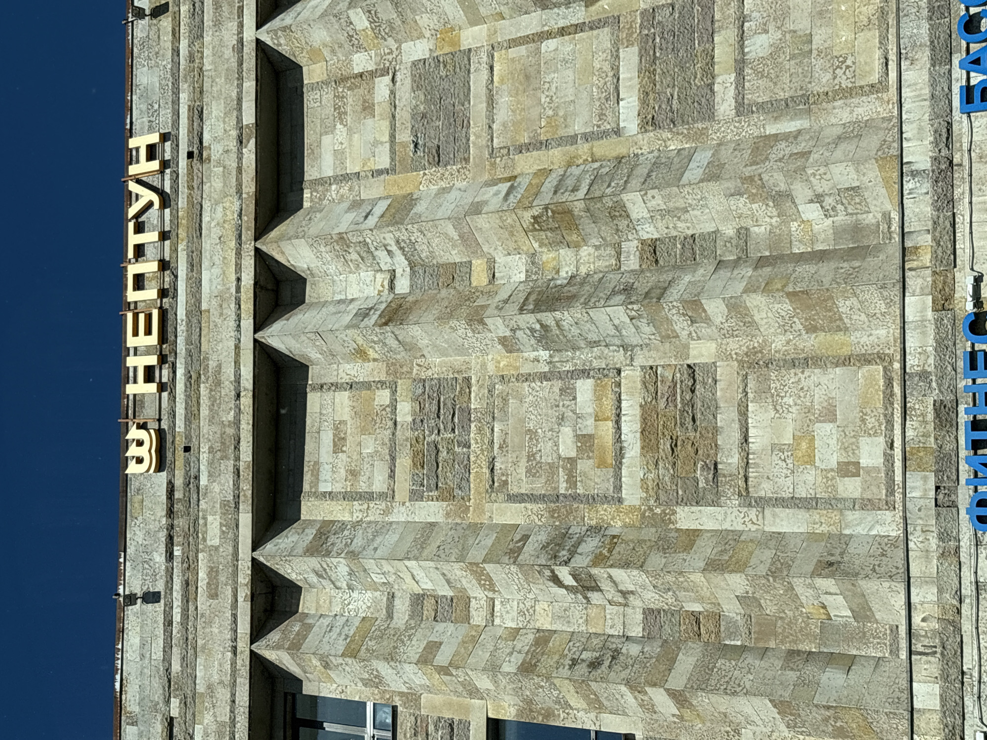 The image shows the facade of a building in Saint Petersburg, Russia, with the golden Cyrillic letters "НЕПТУН" (Neptune) prominently displayed at the top. The building's exterior is constructed of large, textured stone blocks in varying shades of beige, grey, and brown, arranged in a pattern of vertical columns and horizontal bands. There are geometric indentations and raised sections on the facade, creating a sense of depth and architectural detail. Two windows with dark frames are visible on the left side of the building. The sky above is a clear, deep blue. Below the "НЕПТУН" sign, a portion of a blue sign with Cyrillic letters is visible, suggesting a commercial establishment. The lower edge of the image shows more of the stone facade and the beginning of a street or sidewalk level.