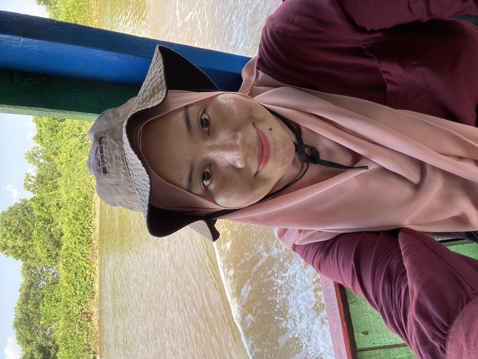 A medium shot selfie captures a woman wearing a maroon long-sleeved shirt, a light pink hijab, and a tan bucket hat with a logo. She is on a boat, with a river and lush green vegetation visible in the background. The boat's railing in the foreground is painted red and green, with blue and green posts visible to the right. The woman is smiling and looking at the camera. The scene appears to be in Banjarmasin, Indonesia, likely on a river or canal.