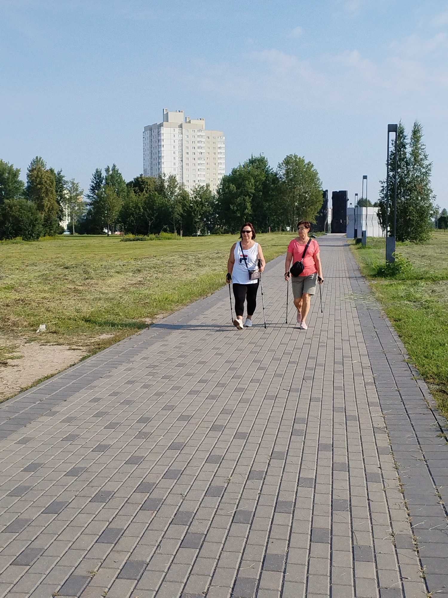 Two women Nordic walking on a paved path in a park. The woman on the left wears a white tank top and black leggings, and the woman on the right wears a pink shirt and khaki shorts. Both women are using Nordic walking poles. The path is lined with grass and trees. In the background, a tall, modern building and several dark, abstract sculptures are visible. The sky is clear and blue. The scene appears to be in a city park. The presence of the Stalin Line memorial complex suggests this image was taken in the vicinity of the memorial, which is located in the Minsk region of Belarus.