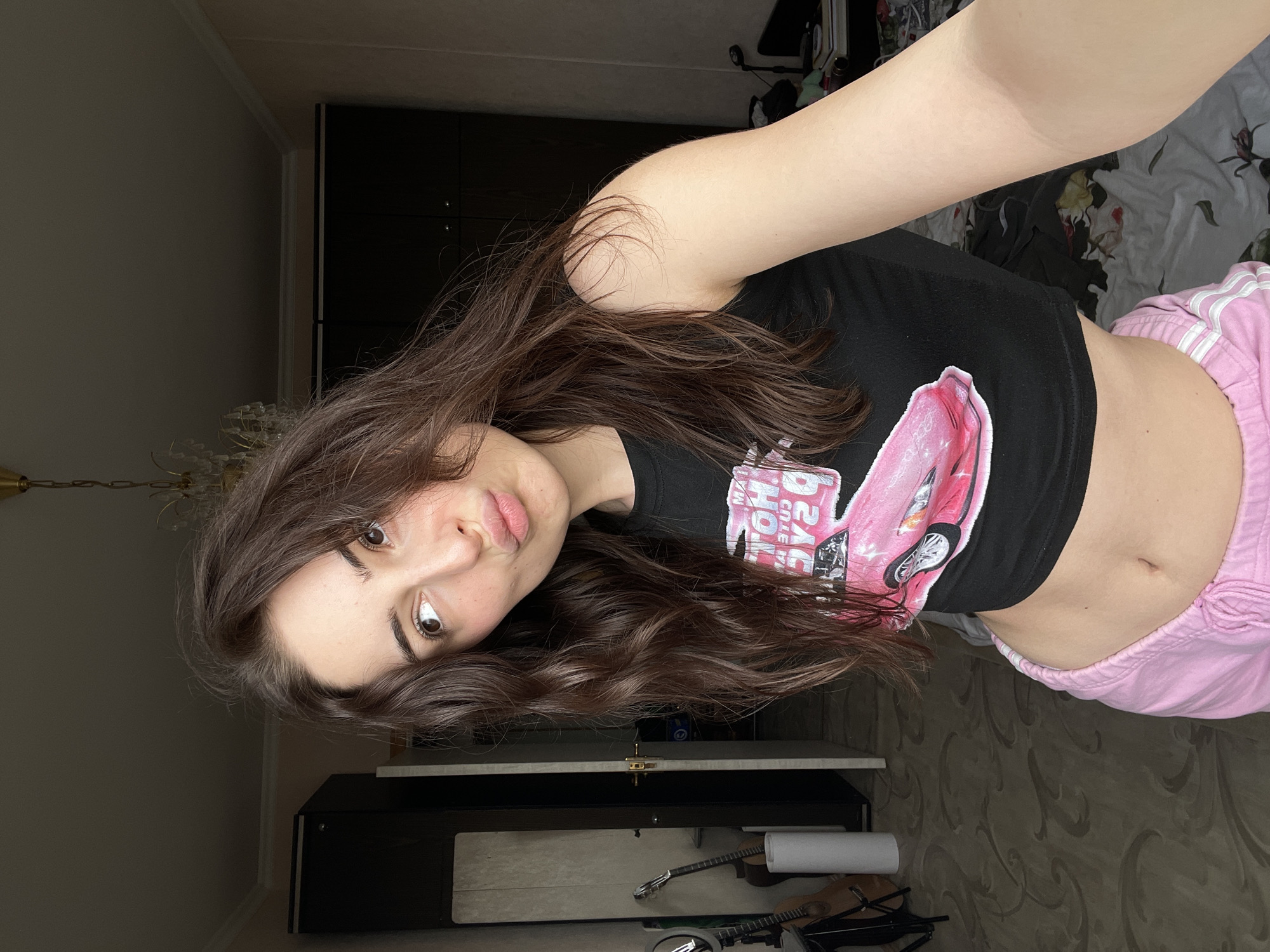 A young woman with long brown hair is posing for a selfie indoors. She is wearing a black crop top with a pink design and pink sweatpants. Behind her, there's dark furniture and a bed. The photo appears to be taken in a residential setting, and there is no indication of the city of Moscow, Russia.