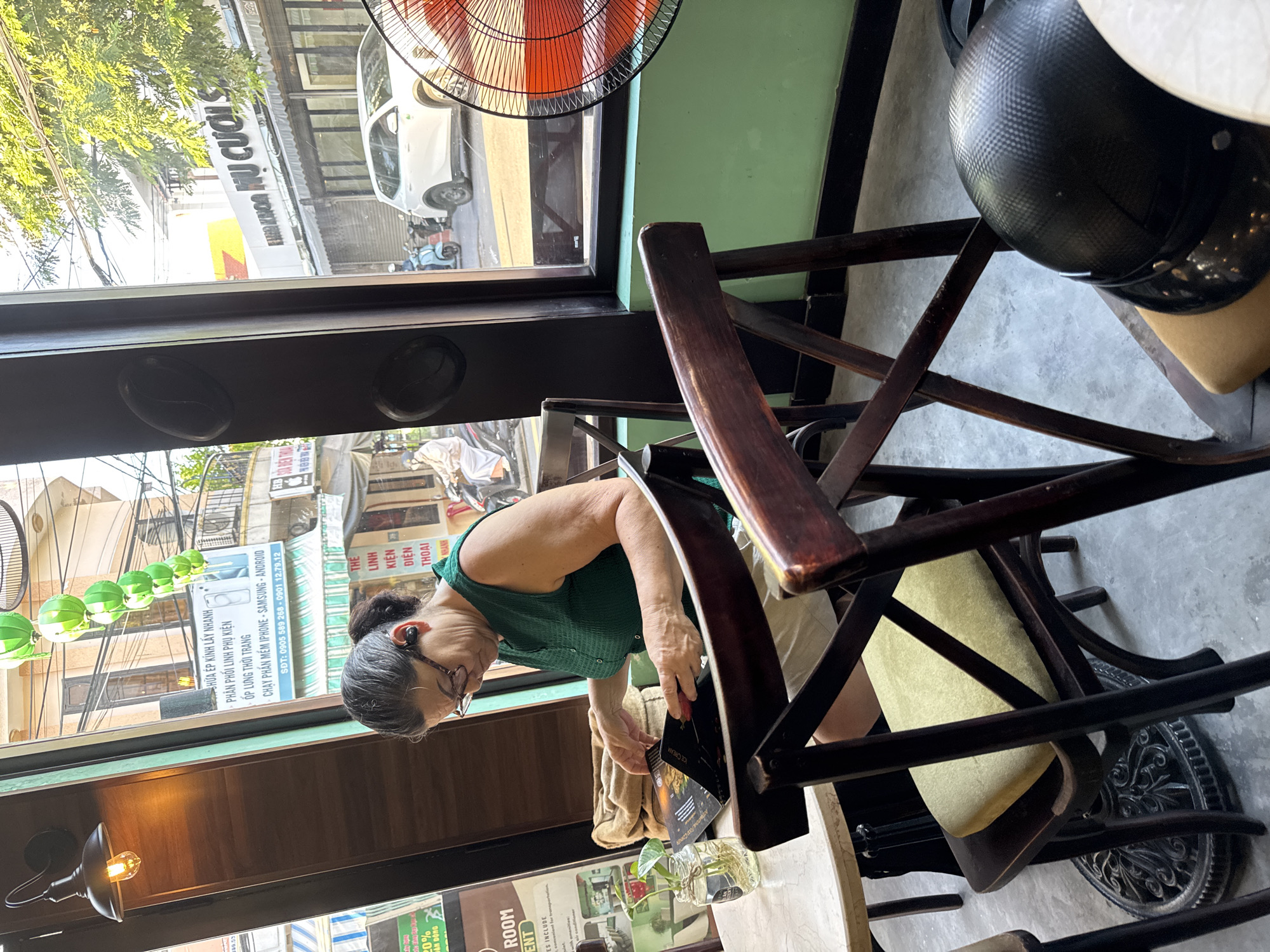 The image shows an older woman with dark hair pulled back and wearing glasses and a green sleeveless shirt, sitting at a table in a cafe in Hoi An, Vietnam. She is looking down at a menu or magazine she is holding open in her hands. There is a glass of water with a plant on the table next to her, and a brown cloth bag behind her. In the background, through a window, street scenes of Hoi An are visible, including buildings, a white SUV, and motorbikes. Several green paper lanterns are strung across the upper portion of the window. Two wooden chairs with light yellow cushions are in the foreground, one occupied by the woman and another empty. A black helmet with a carbon fiber pattern rests on the empty chair in the lower right corner. The cafe appears to have a rustic ambiance with dark wooden furniture and light green walls.