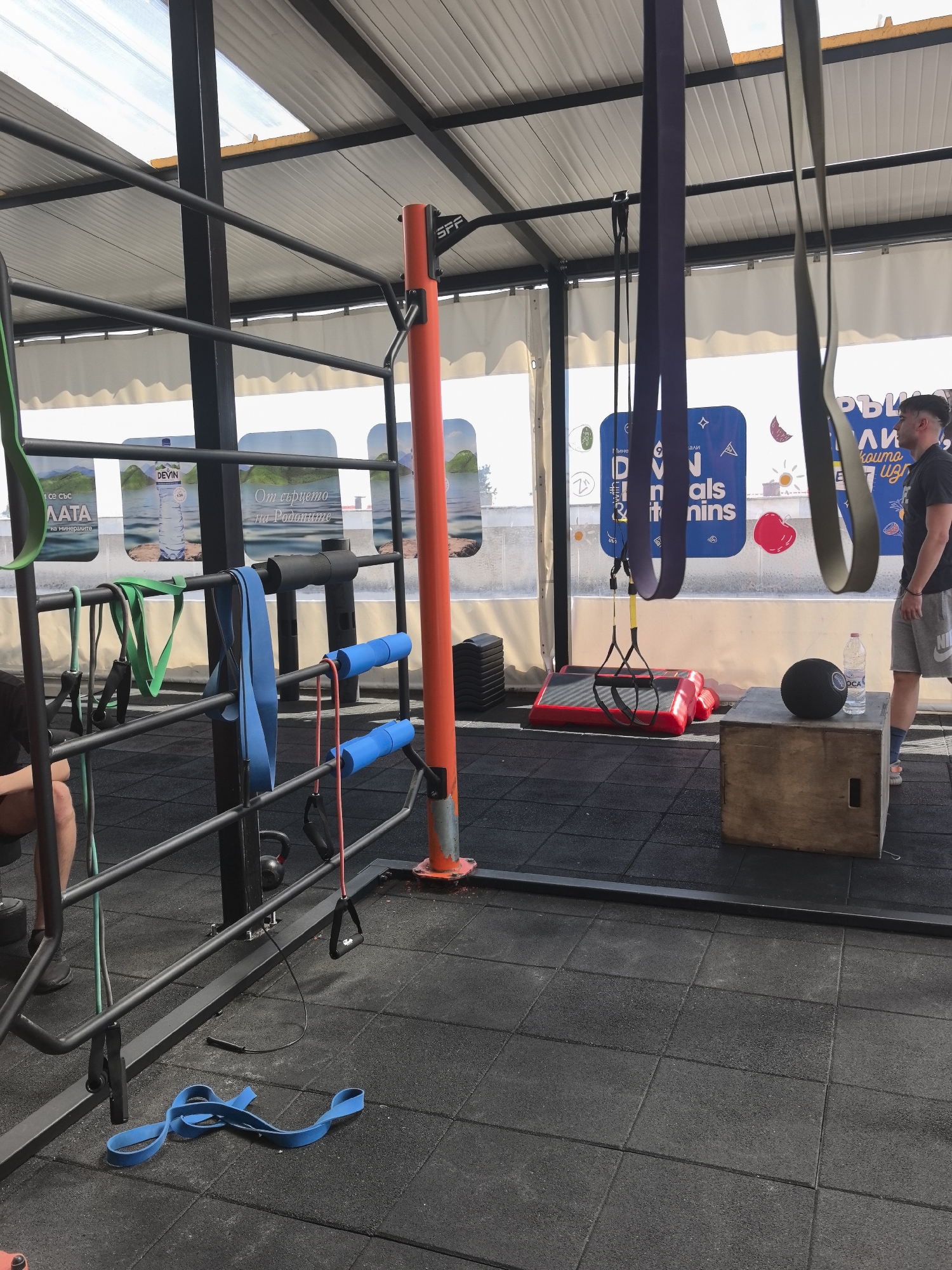 Here is a description of the image:
**Location:** The image appears to be taken at an outdoor gym facility located in Stara Zagora, Bulgaria. This is deduced from the signage and the "DEVIN" bottled water visible in the background.
**Scene:** The image captures an outdoor workout area with a variety of exercise equipment. The gym is covered by a translucent roof, and the walls are partially enclosed with advertising signage.
**Objects:**
* Various exercise equipment including: pull-up bars, resistance bands, suspension trainers (TRX), plyometric boxes, aerobic steps, medicine balls.
* A bottle of "OCA" water and another of "DEVIN" water are visible.
* Rubber flooring that is likely gym matting.
**People:**
* A man is standing on the right side of the frame, wearing a dark t-shirt and light gray athletic shorts. His attention seems to be directed towards something outside the frame.
* Part of another person, including part of a leg and forearm, is visible to the left of the frame.
**Environment:**
* The gym is outdoors, but under a sheltered structure. The lighting is bright, suggesting it is daytime.
Overall, the image is of a well-equipped outdoor gym facility in Stara Zagora, Bulgaria.