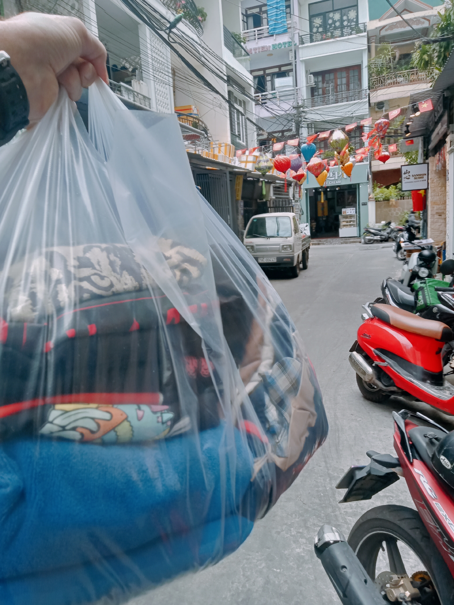 A person holds a large, clear plastic bag filled with items including what appears to be blankets and clothing in a narrow street in Nha Trang Ward, Vietnam. The street is lined with buildings, some decorated with strings of lanterns and flags. Parked motorbikes are visible along the side of the street. A small white truck is parked further down the street in front of a shop. Overhead, tangled electrical wires span the street. The street appears to be paved and relatively clean. The focus is on the bag and its contents, with the street scene providing the background. The time of day appears to be daylight, with ambient lighting.