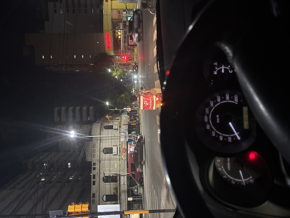 A night shot from inside a car, focusing on the dashboard with illuminated gauges. Outside, city streets are visible, lit by streetlights, illuminating buildings of various heights. A delivery person with a large "PedidosYa" bag is standing on the crosswalk. Street signs and traffic lights are also present. The overall scene depicts a typical urban night.