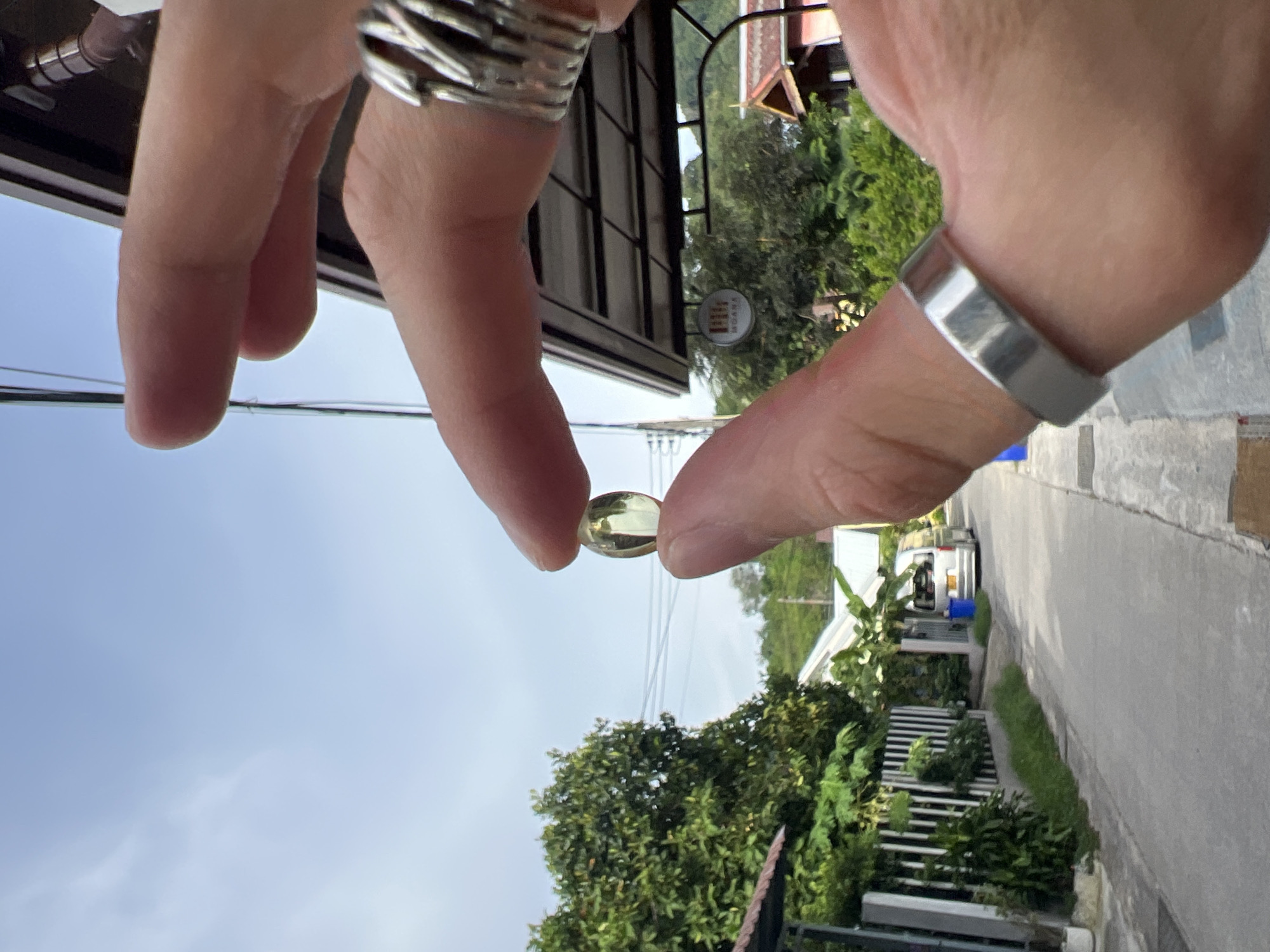 A close-up shot focuses on a person's hands holding a translucent, oval-shaped pill between their thumb and index finger. The pill is pale yellow and appears to be a supplement capsule. The person is wearing two silver rings: one intricate, multi-banded ring on the index finger and a simple, wide band on the ring finger. The background reveals a street scene in what appears to be Choeng Thale, Thailand. Lush green trees line the left side of a paved road, bordered by a white fence. A silver van is parked on the right side of the road, with a blue trash bin nearby. In the distance, traditional Thai-style buildings with reddish-brown roofs are visible, nestled amongst more foliage. The sky above is a hazy, light blue with faint clouds. The overall lighting suggests it is daytime, possibly late morning or early afternoon.
