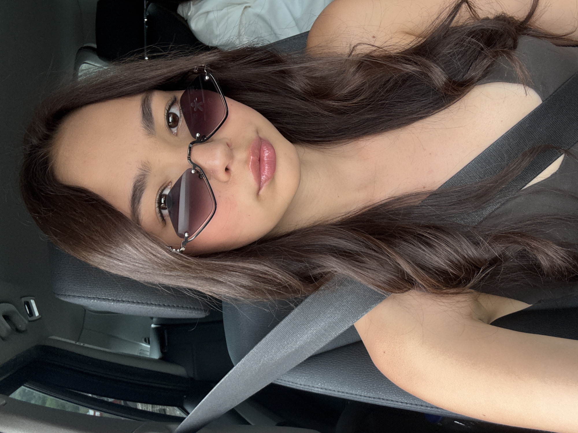 A woman with long brown hair is in the passenger seat of a car. She wears sunglasses and a dark-colored top, and a seatbelt is fastened across her chest. The car interior is visible, including the leather seat and dashboard. The specific location within the city of Unknown, Unknown cannot be determined from the image.