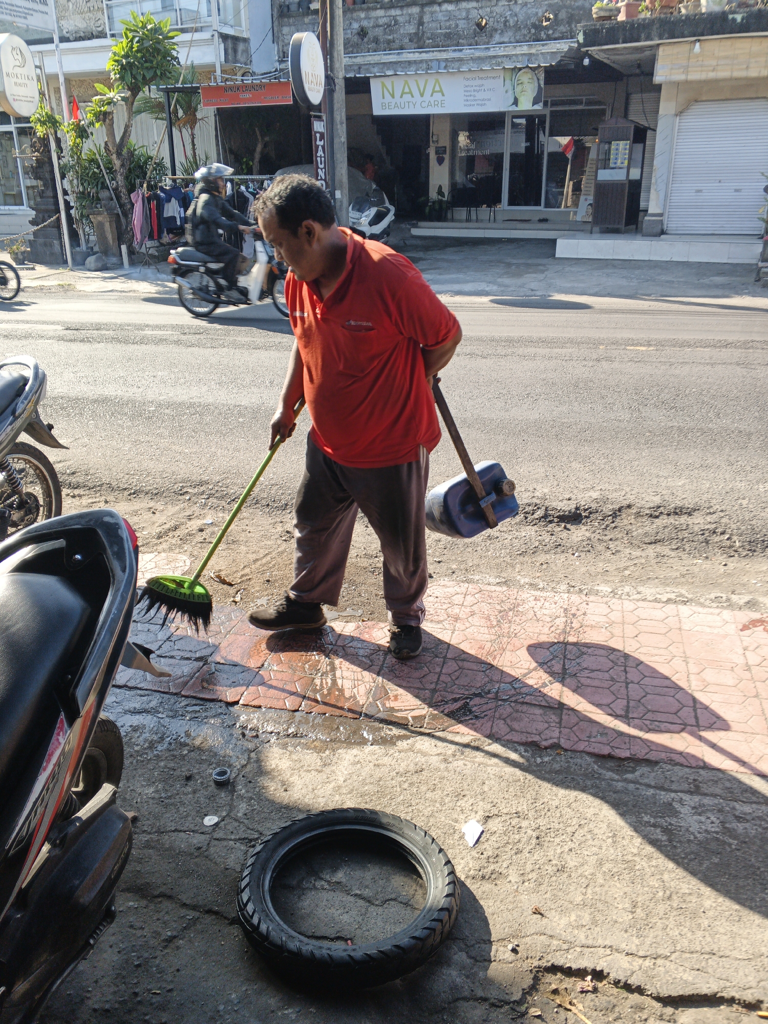 A man wearing a red shirt and gray pants is sweeping the sidewalk in front of shops in Sukawati, Indonesia. He is holding a green broom in his right hand and a blue plastic container on a wooden handle in his left. The sidewalk is made of red patterned tiles and is wet in some areas. Next to the sidewalk is a street with a motorcycle parked on the left side of the frame. Behind the man, there are several shops with signs and awnings. One sign reads "Nava Beauty Care". The scene is bathed in bright sunlight, casting long shadows on the ground. A discarded tire lies on the cracked concrete next to the motorcycle.