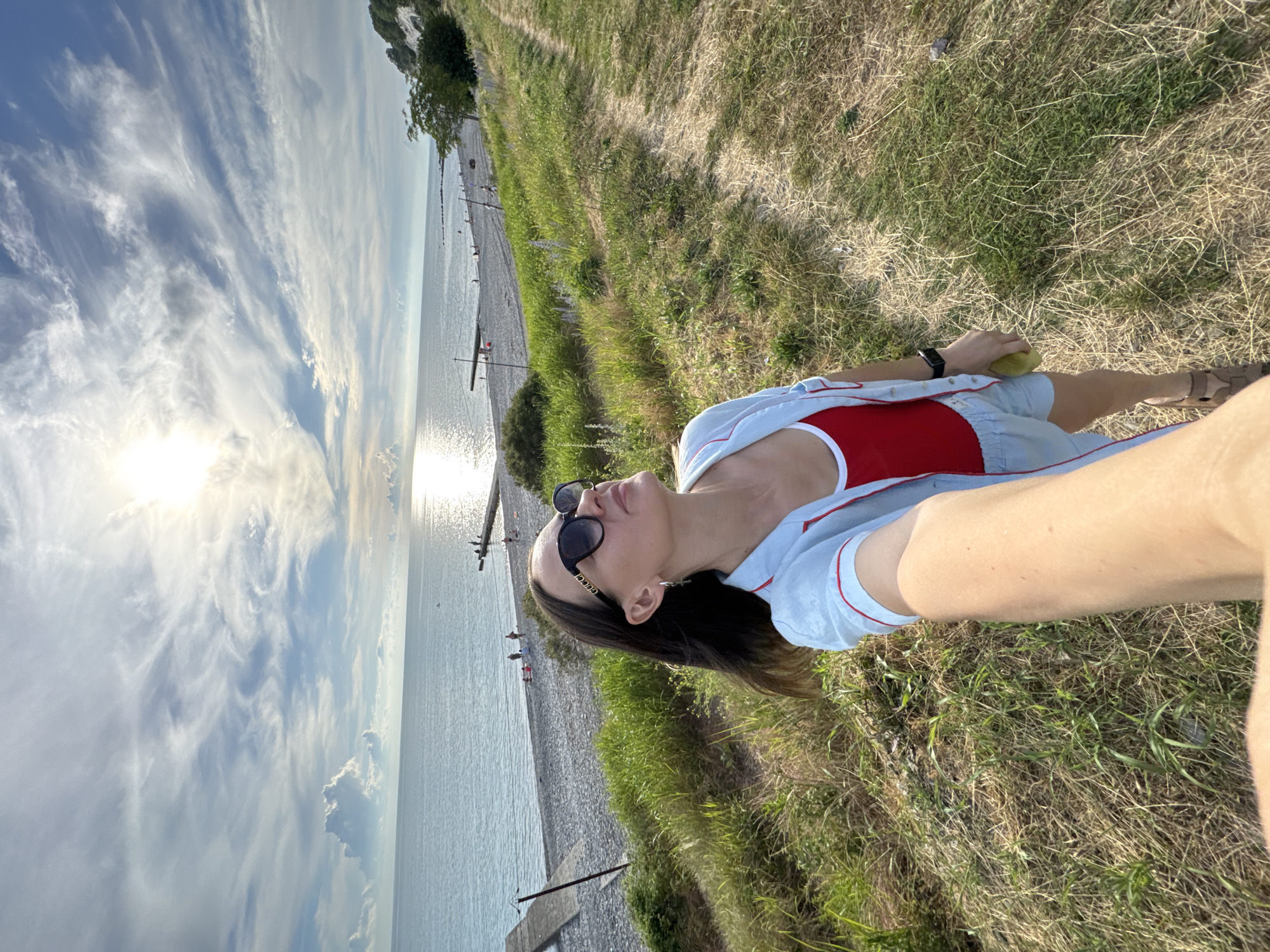 A woman takes a selfie on a cliff overlooking a pebble beach and the Black Sea in Sochi, Russia. The woman is wearing sunglasses, a red swimsuit, and light blue shorts and a matching shirt. She is holding a pear in her right hand. The sun is bright and reflected on the water. Several people are visible on the beach, and a pier extends into the sea. The sky is partly cloudy with scattered cumulus clouds. The ground is covered in dry grass.