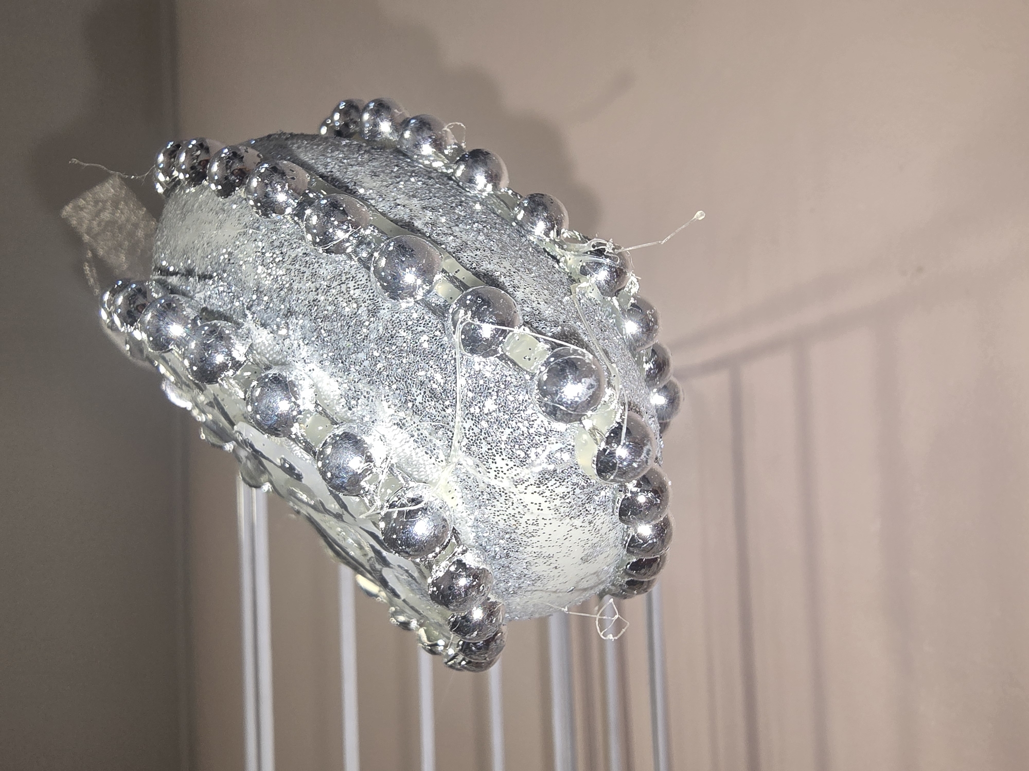 The image shows a close-up, low-angle shot of a silver, glitter-covered Christmas ornament hanging from a string. The ornament is oval-shaped and adorned with a string of small, silver, beaded balls around its circumference. Some clear adhesive is visible holding the beaded string in place. The background is out of focus, showing horizontal lines that appear to be part of a drying rack or similar structure. A soft, pinkish-tan surface is visible below the lines. There is no indication of any specific location in Ulan-Ude, Russia.