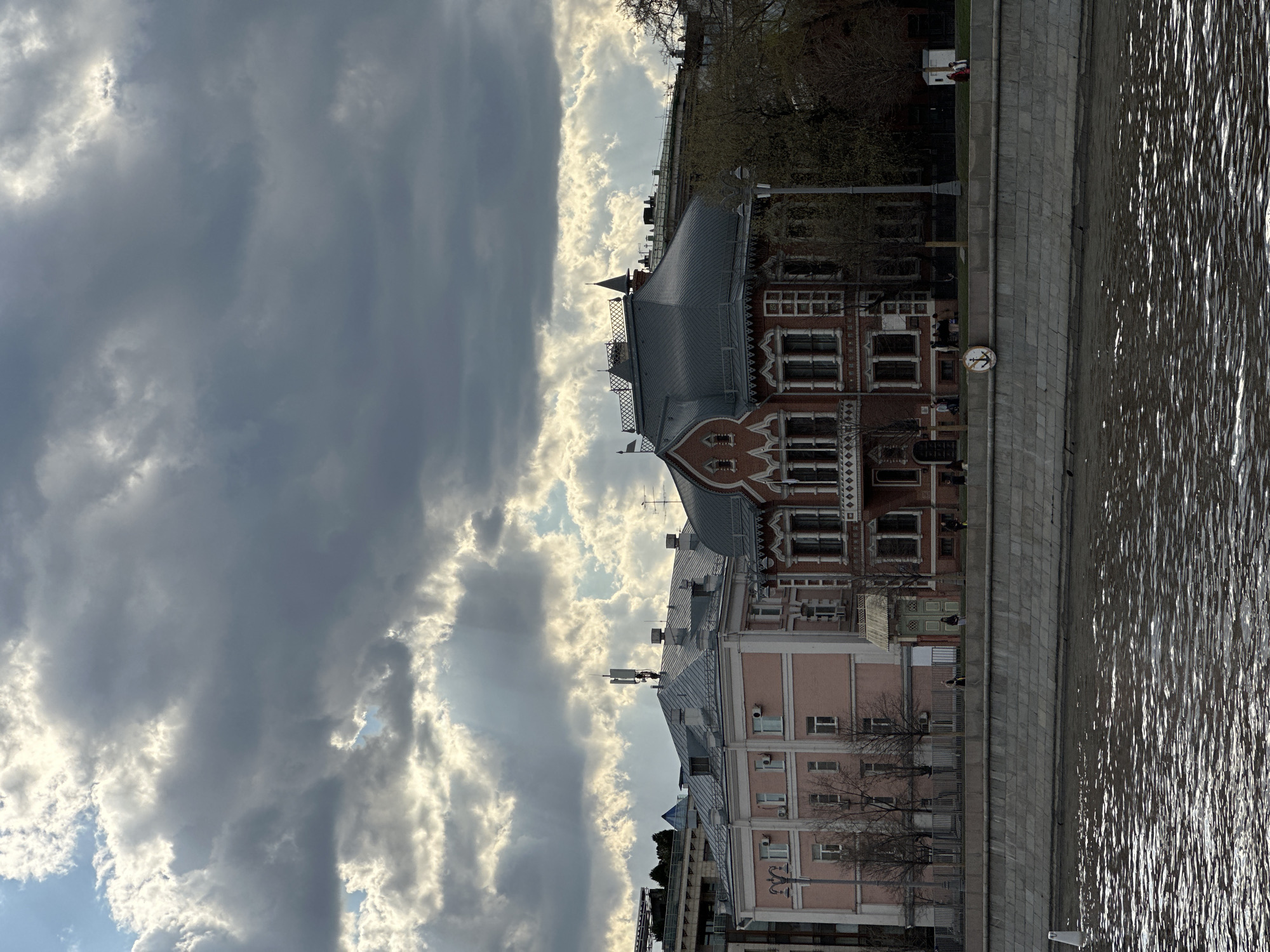 The image captures a view of the embankment in Moscow, Russia, on a cloudy day. The water of the Moscow River is visible in the foreground, reflecting the light. Adjacent to the embankment is a stone walkway, lined with buildings. The most prominent building is a red-brick structure with white decorative trim and a multi-tiered roof. A lighter-colored building is connected to it. The sky above is filled with dark, dramatic clouds, with patches of bright sunlight breaking through.
