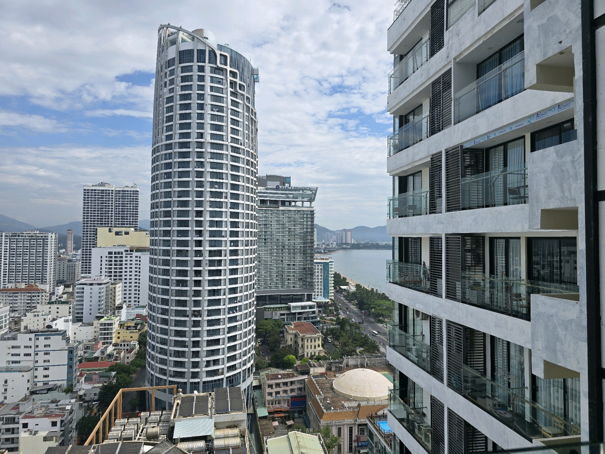 The image captures a panoramic view of Nha Trang, Vietnam, looking out towards the coast. In the foreground, a modern white building with multiple balconies overlooks the scene. To the left, a tall, curved skyscraper with a grid-like facade dominates the skyline. Behind it, other buildings of varying heights and architectural styles are visible, including a prominent glass-fronted building. The middle ground shows a coastal road with traffic, fringed by palm trees and leading to a beach with a few people. The ocean stretches to the horizon, meeting a hazy sky with scattered clouds. The overall impression is one of a bustling, modern city situated along a picturesque coastline.