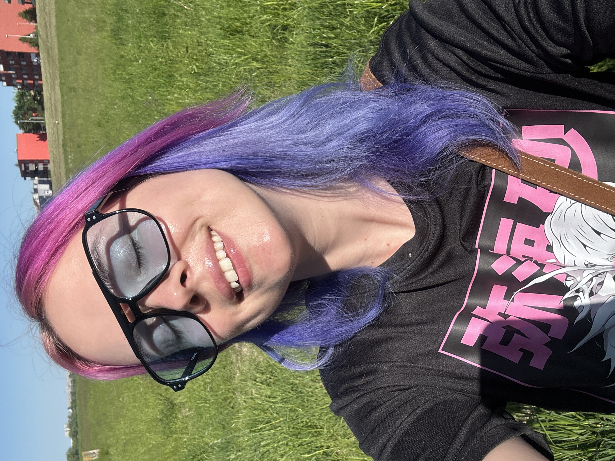 A fair-skinned woman with pink and purple hair is lying on a grassy field in the City of Zagreb, Croatia. She is wearing black-rimmed glasses and a black t-shirt with a pink graphic design. A brown strap crosses her body. In the background, there are buildings and a clear blue sky.