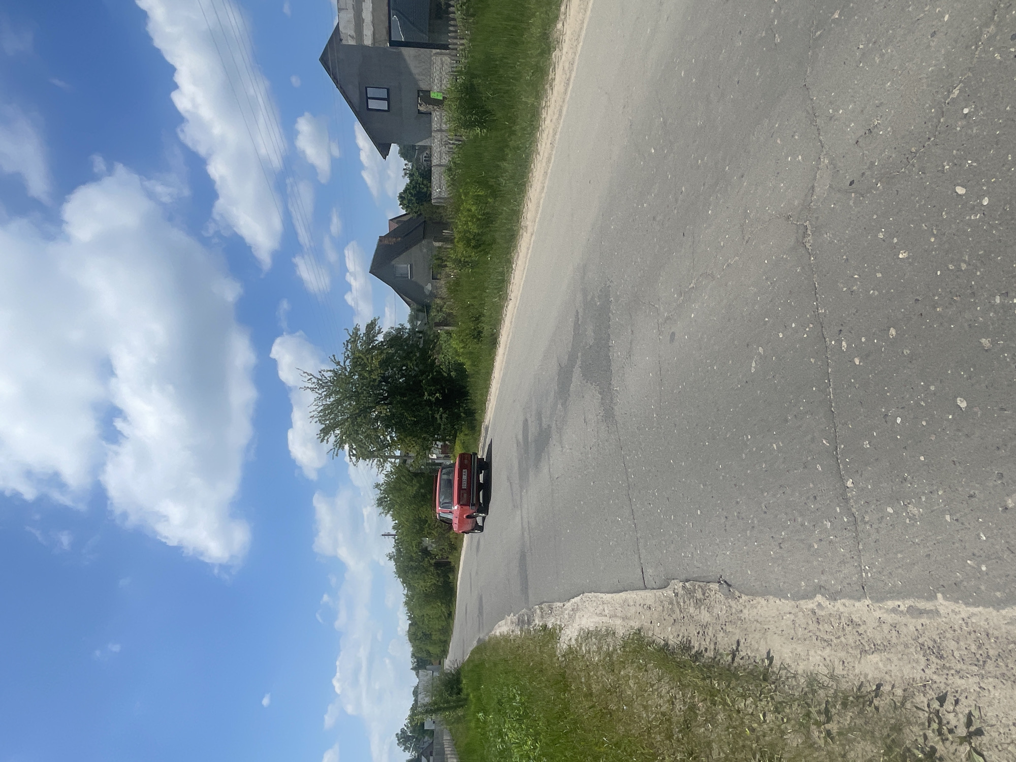 A bright red car is driving down a paved road in a rural area with houses and trees on either side. The sky is blue with white clouds. The road is in fair condition with some cracks and patches. The car appears to be a Soviet-era model. There is grass and vegetation growing along the side of the road. The houses appear to be under construction or recently built. The overall scene suggests a quiet, sunny day in a village or small town.