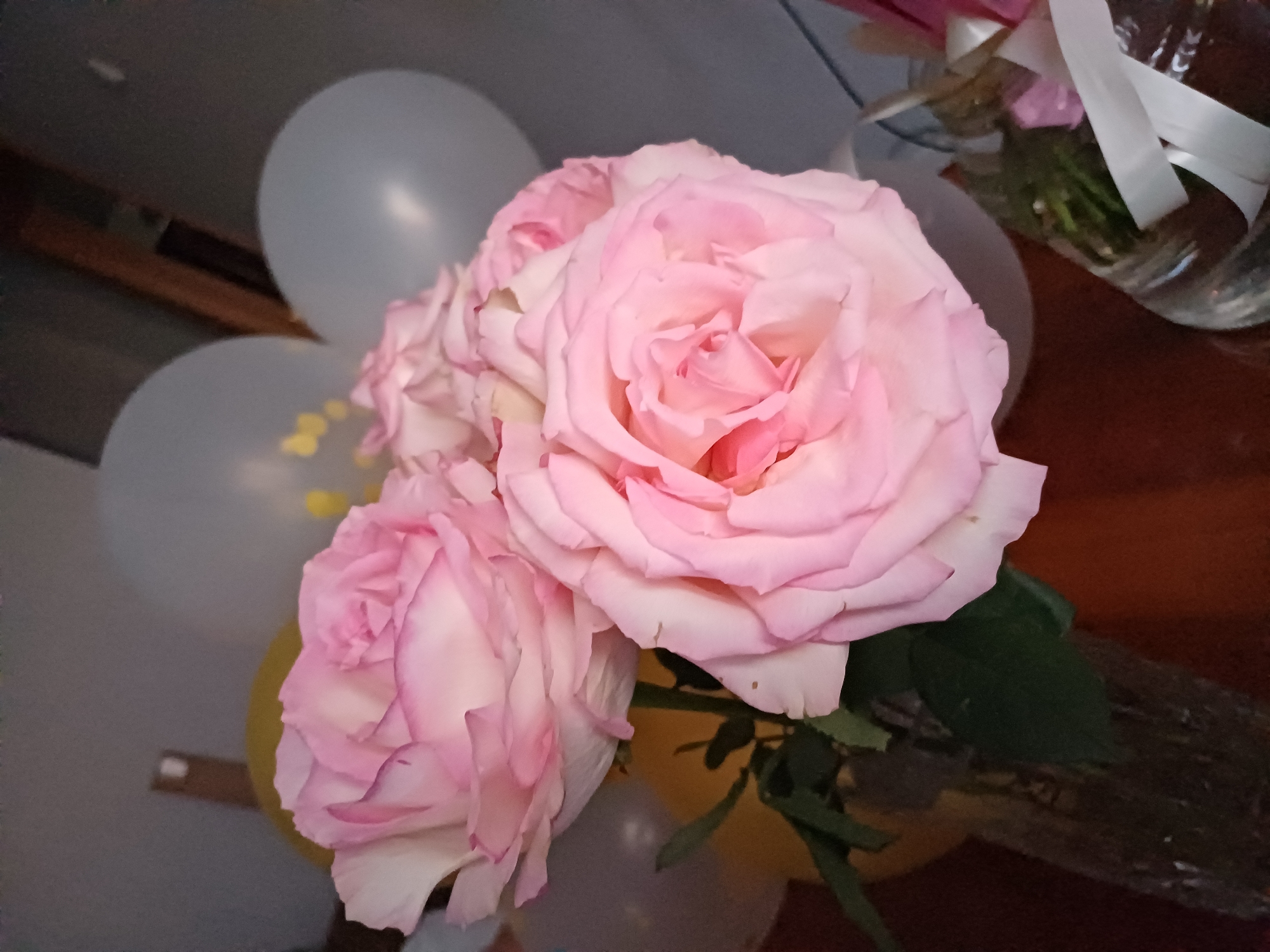 The image shows a close-up view of a bouquet of pink roses in a clear vase. The roses are large and fully bloomed, with soft pink petals that are lighter towards the center. Behind the roses, several light gray and yellow balloons are visible. The background is out of focus but appears to be an indoor setting with a dark wall and wooden elements. The lighting is somewhat dim, casting soft shadows.