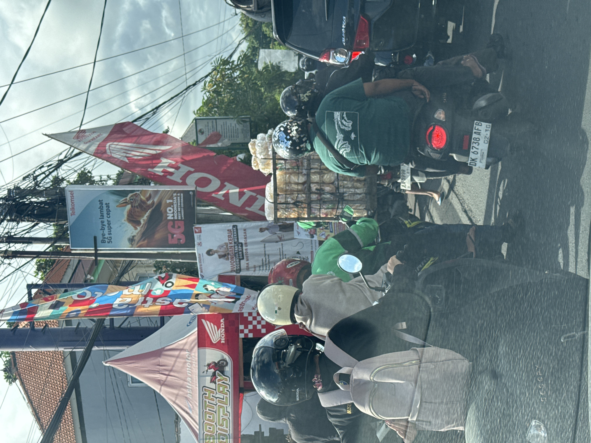 The image depicts a congested street scene in Kuta Selatan, Indonesia, as seen from inside a vehicle. The view is obstructed by the vehicle's windshield. Several motorcycles are visible, lined up in traffic. One motorcycle has a large quantity of what appears to be packaged snacks strapped to the back. Riders are wearing helmets; one has a green shirt with a white design on the back, and another wears a helmet with a black and white patterned design. Street signage and banners are present, advertising various businesses and services, including Astra Motor, Honda, Telkomsel, and job openings. Power lines crisscross overhead. The license plate on a motorbike reads "DK 6738 AFB." The sky is overcast.