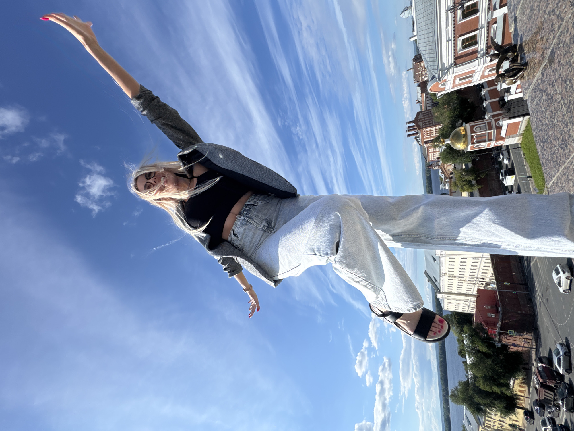 The image depicts a woman with blonde hair and glasses posing with her arms outstretched against a bright blue sky with wispy white clouds. She is wearing a black crop top, a grey blazer, and light-wash jeans. Her left leg is bent and extended forward, and she is wearing black sandals. In the background, a cityscape is visible, featuring red-brick buildings with white trim, a golden-domed structure, trees, a river, and a street with cars. The perspective is low-angle, looking up at the woman. The location is identified as Novokuybyshevsk, Russia.