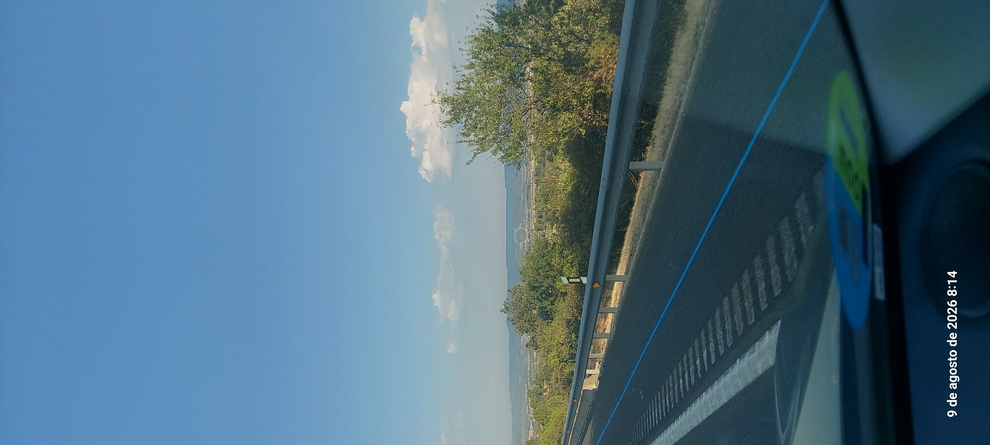 The image depicts a roadside view on a clear, sunny day. The foreground shows a paved road with white lane markings and a distinctive blue line running along the edge of the road. A metal guardrail runs parallel to the road, separating it from a grassy verge. Beyond the verge, a landscape of rolling hills covered in green vegetation and scattered trees stretches into the distance. A large transmission tower with a hexagonal frame is visible in the middle ground. The sky is a vibrant blue with a few scattered white clouds. The timestamp in the lower left corner indicates the image was taken on August 9, 2026, at 8:14 AM. The location is identified as Prenafeta, Spain.
