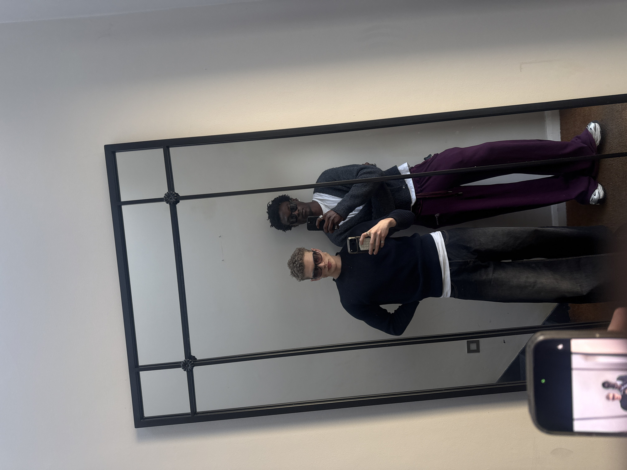 A mirror on a white wall reflects two people taking a selfie. The person closest to the camera in the mirror wears a black sweater, sunglasses, and dark pants. The other person in the mirror wears a white t-shirt, a gray cardigan, sunglasses, and purple pants. A phone is in the foreground, presumably taking the photo of the mirror's reflection. It is unclear whether the location is in London, United Kingdom.