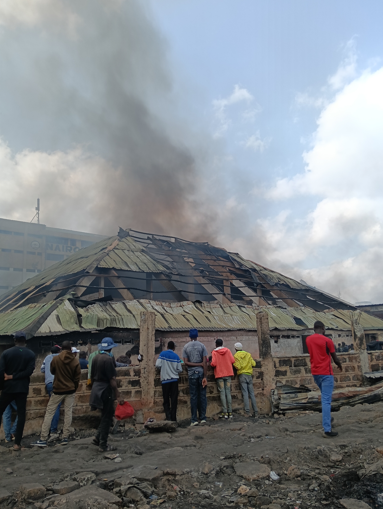In Nairobi, Kenya, a building with a corrugated iron roof is engulfed in thick black smoke, indicating a fire. The roof is partially collapsed, revealing the wooden structure beneath. Several people are gathered, observing the scene from a distance. Some stand in groups, while others are more spread out. The ground in front of the building is littered with debris, suggesting damage or destruction. In the background, a multi-story building with the partial inscription "NAIROBI" is visible, indicating the urban setting. The sky is partly cloudy.