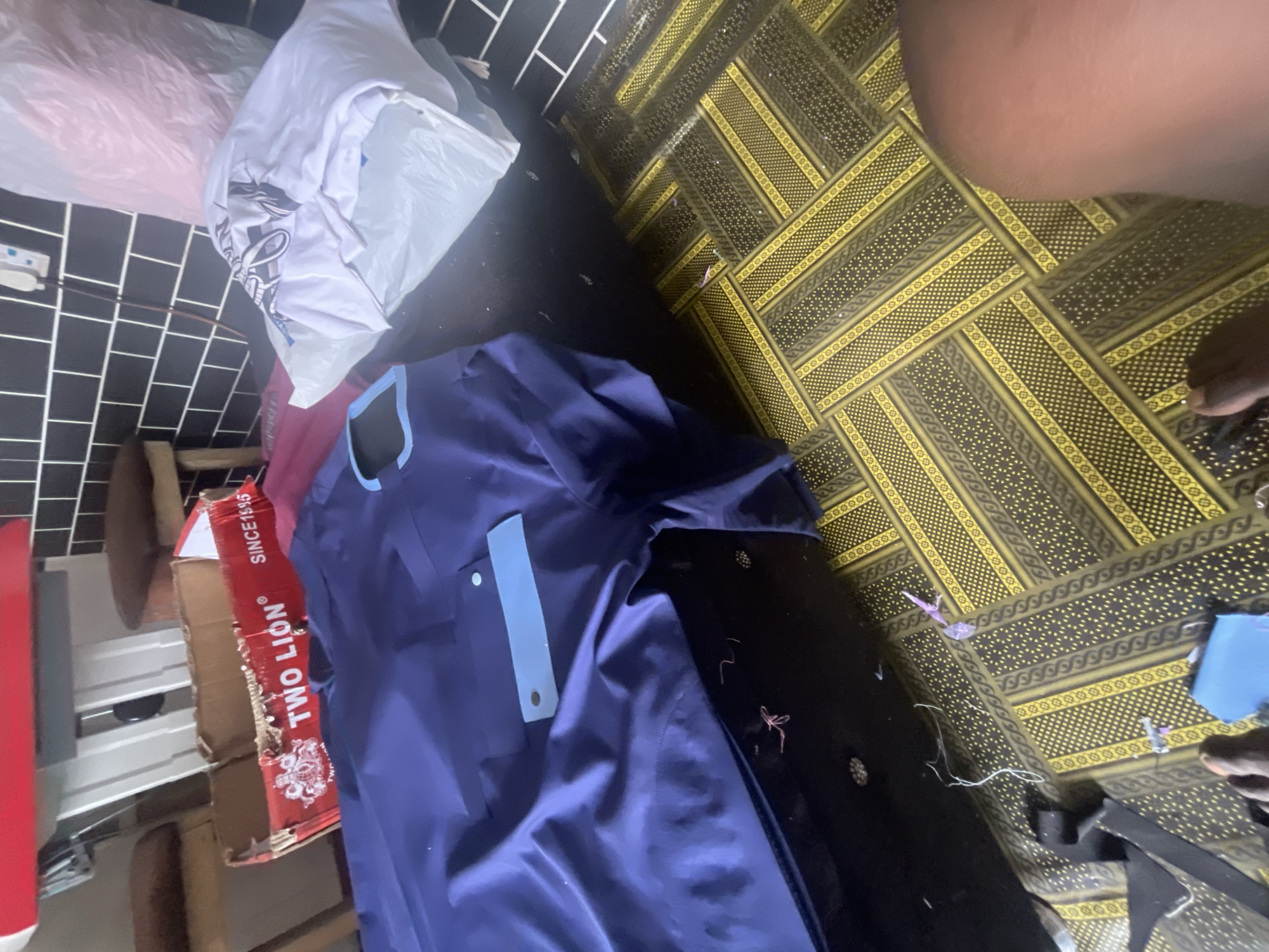 In Jauro Yinu, Nigeria, a scene unfolds within a domestic setting, characterized by a utilitarian aesthetic. The foreground is dominated by a blue garment, possibly a traditional African attire, laid out on a dark surface. Its rich color and fabric suggest it's ready for wear or has recently been unpacked. Adjacent to it lies a white t-shirt, neatly folded, indicating a sense of order amidst the clutter.
The background reveals a juxtaposition of textures and colors. Black and white patterned tiles line the walls, offering a modern contrast to the rustic, red cardboard box situated on top of the garment. The box, emblazoned with the "TWO LION" logo and "SINCE 1985", hints at the origin of the items or perhaps a recent delivery. Two wooden stools, their surfaces worn with time, stand as silent witnesses to past activities.
The floor, a vibrant mosaic of yellow and dark brown geometric patterns, adds a lively touch to the otherwise subdued environment. Scattered amidst the patterns are small, unidentified objects, possibly remnants of packaging or daily activities. The presence of a pink plastic bag, partially visible, suggests a recent acquisition or disposal.
The scene is devoid of human presence, yet the arrangement of objects implies human activity and domestic life. The lighting, though not explicitly detailed, appears to be a mix of natural and artificial sources, casting subtle shadows and highlighting the textures of the various items. The overall impression is one of a lived-in space, where practicality and personal belongings converge.