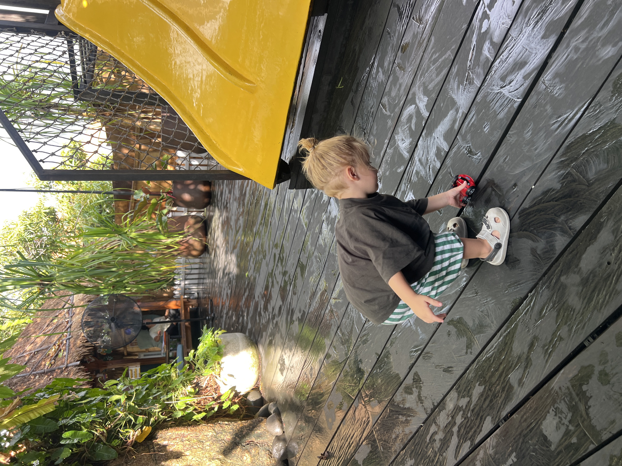 A young child with blonde hair in a bun, wearing a dark t-shirt and green and white striped shorts, crouches on a wet wooden deck, playing with a small red toy car. The child is looking down at the toy. Behind the child, a bright yellow slide curves upwards, partially obscured by a black metal fence with netting. In the background, lush green foliage and a thatched-roof structure are visible, suggesting an outdoor tropical setting. The wooden deck is dark and appears to have been recently rained on, with reflections of light and greenery visible on its surface. This scene is set in North Kuta, Indonesia, likely in a resort or playground area.