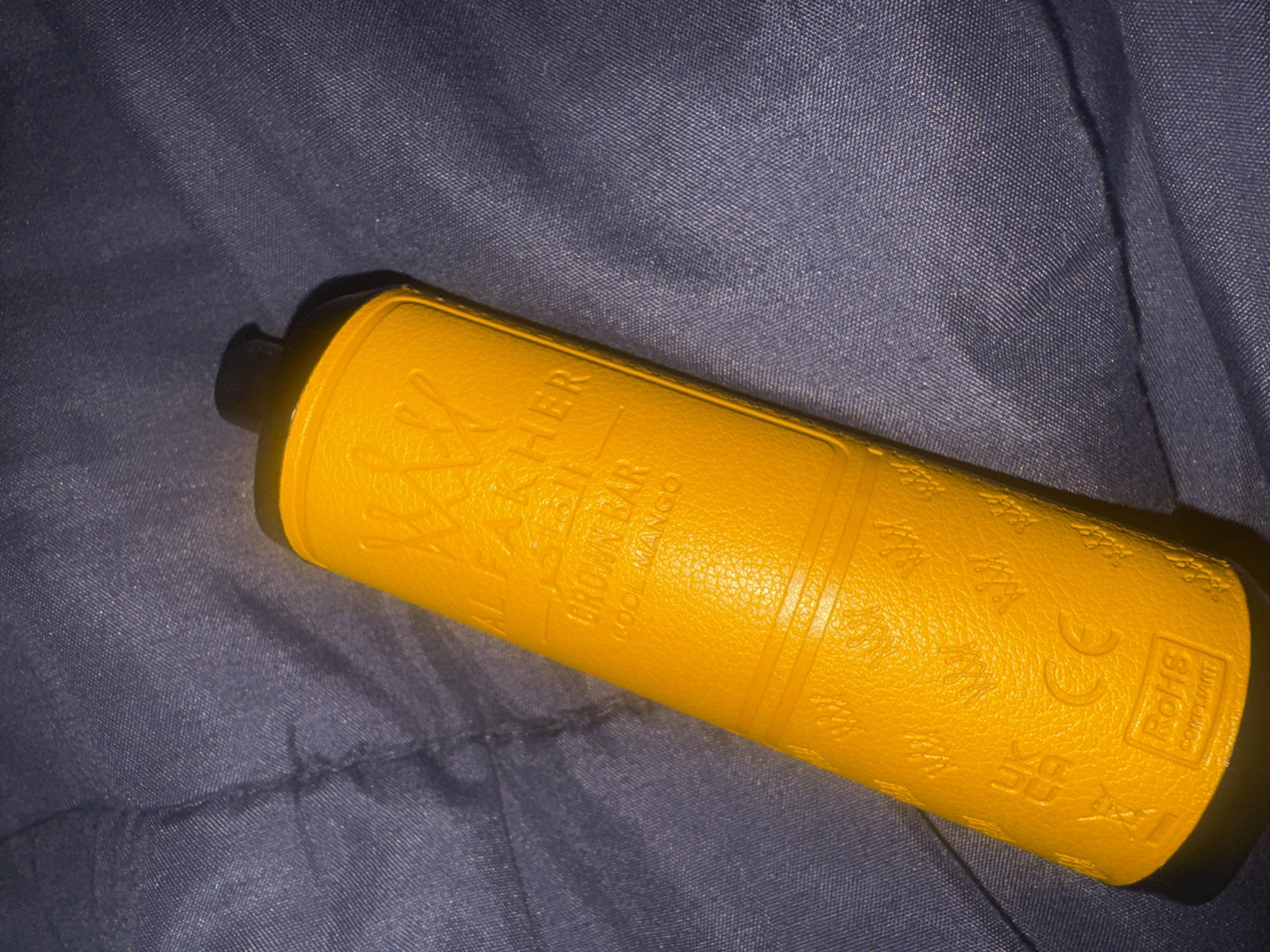 A yellow disposable vape pen rests on a dark blue textured surface, likely fabric. The vape pen has embossed text and symbols. The top portion of the pen is obscured by a black cap. Text visible on the vape pen includes "AL FAKHER," "CROWN BAR," and "COOL MANGO." Near the bottom, there are markings including "UK CA," a "CE" symbol, and a "RoHS COMPLIANT" stamp. The background is a dark blue fabric with visible creases and a subtle pattern. There are no people or other objects in the frame. The image is taken from a slightly angled overhead perspective. The location of this image within the city of Abuja, Nigeria, cannot be determined from the visual information provided.