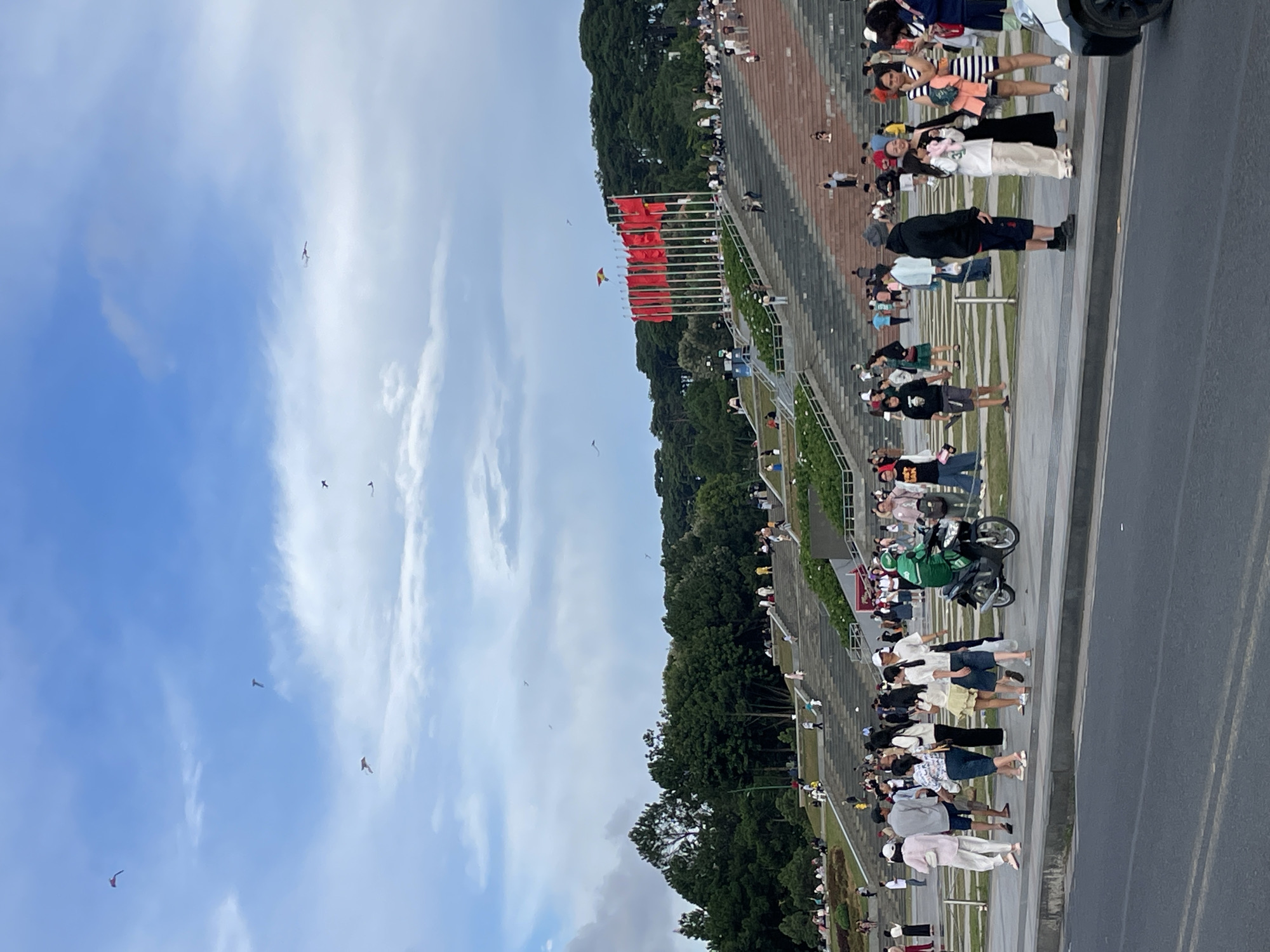 This is a vertical, full-shot photograph taken outdoors, possibly in Da Lat, Vietnam, under a bright, cloudy sky. The scene captures a gathering of people, many of whom are seen flying kites in the vast blue expanse. The foreground is dominated by a paved area bordering a large set of tiered, red-brown steps that ascend towards a densely wooded hillside.
The people are scattered across the paved area and on the steps, appearing relaxed and engaged in various activities. Some are standing, others are sitting or walking, and a few are actively flying kites. The kites are of various colors and shapes, adding dynamic elements to the sky. The overall atmosphere seems festive and casual, suggesting a public event or a popular recreational spot.
A motor scooter with a green bag is parked on the paved area, indicating possible transportation for some attendees. The time of day appears to be late afternoon or early evening, given the quality of the light and the long shadows cast. The weather seems pleasant, with a mix of sun and clouds, allowing for outdoor activities like kite flying.
The presence of numerous people suggests this is a well-frequented location. The red-brown steps might be part of a monument, amphitheater, or a large public park designed for gatherings. The lush green trees on the hillside provide a natural backdrop to the scene. No visible text is present in the image.