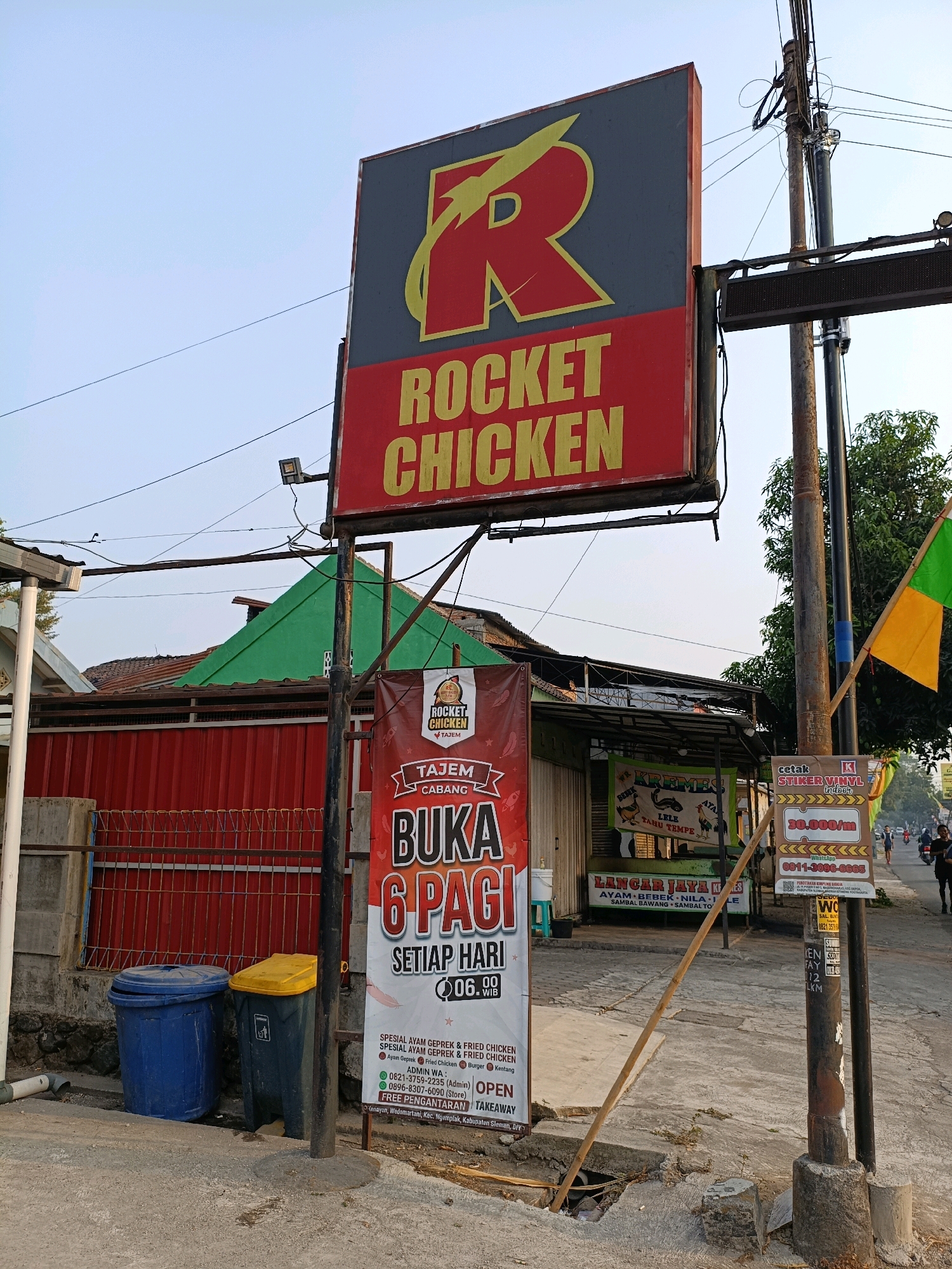 The image shows a street scene in Ngemplak, Indonesia, featuring a prominent "Rocket Chicken" restaurant sign. The sign is large and red with yellow lettering, displaying the restaurant's name and a stylized "R" logo. Below the main sign, a vertical banner advertises "Rocket Chicken Tajem Cabang" with hours of operation from 06:00 to 00:00 WIB and "FREE PENGANTARAN" (free delivery). Other signs visible include one for "KREMEZ" offering "LELE" and "TAHU TEMPE," and another for "LANGAR JAYA KEBAB" advertising "AYAM BEBEK NILA" and various sambals. A utility pole stands to the right of the signs, with wires and a small LED display attached. Two trash bins, one blue and one yellow, are located to the left of the banners. In the background, there are buildings with red and green roofs, a fence, and a tree. Several people are walking and riding motorbikes on the street in the distance. The overall environment appears to be a commercial area with businesses lining the road.