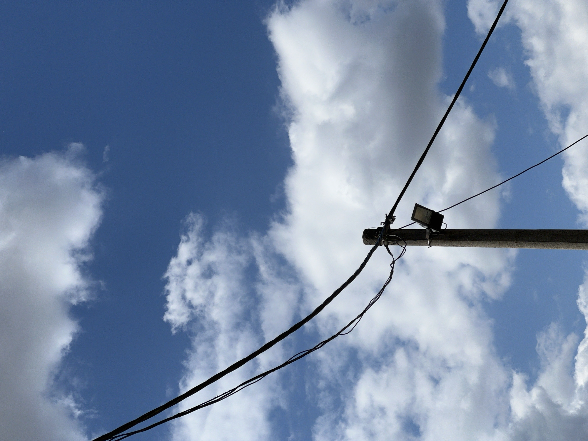 The image depicts a concrete utility pole in Lanžhot, Czechia, against a backdrop of a blue sky with white, puffy clouds. Two thick black electrical wires run diagonally across the frame, connected to the top of the pole. A smaller, darker wire is also visible, attached to the pole lower down. Mounted on the pole, below the main wires, is a rectangular light fixture. The overall scene is a typical outdoor view of urban infrastructure.
