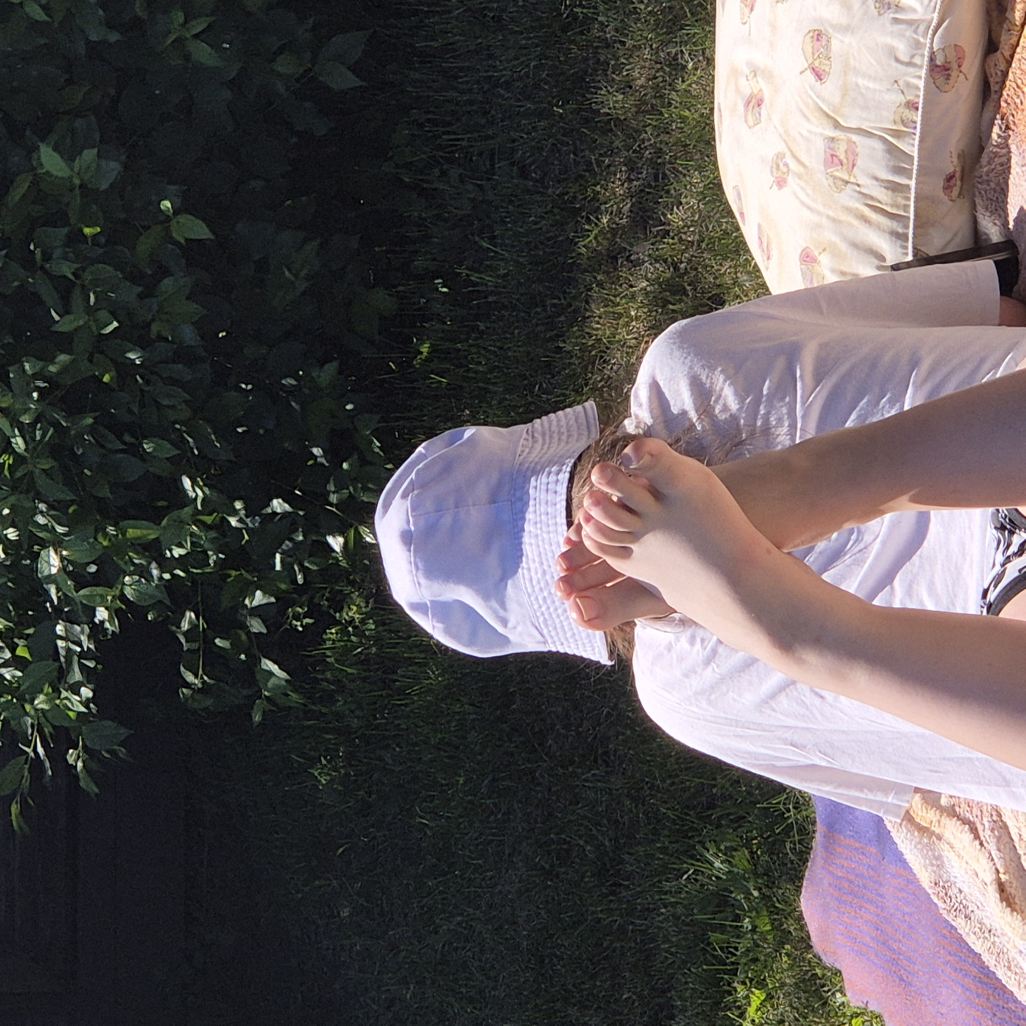 A person is lying on a blanket outdoors, wearing a white t-shirt and a white bucket hat. Their legs are raised and crossed, with their feet positioned in front of the hat. A patterned pillow is placed to the person's right. The background consists of green grass and dense foliage. The scene appears to be set in a residential area, possibly a backyard in Новостройка, Russia.