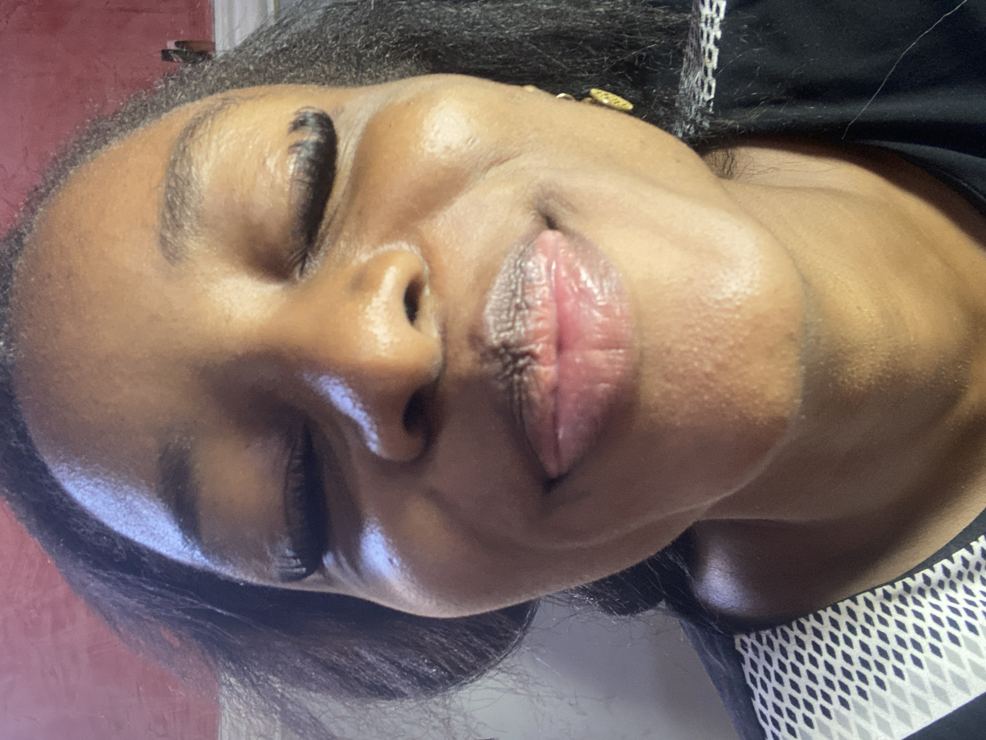 A close-up, head-on shot features a Black woman with her eyes closed and tongue sticking out. She has long, dark hair that is partially visible framing her face. The woman is wearing makeup, including mascara and eyeliner, and has long eyelashes. Her lips are slightly parted, revealing her tongue. A gold earring is visible on her right ear. She is wearing a black and white patterned garment, possibly a shirt or dress, with a mesh-like design around the collar area. The background is a reddish-pink wall with some texture. The lighting suggests an indoor setting, potentially a home, given the informal pose and close-up framing of the subject. The image lacks any distinct geographical markers to indicate it was taken in Jos, Nigeria.